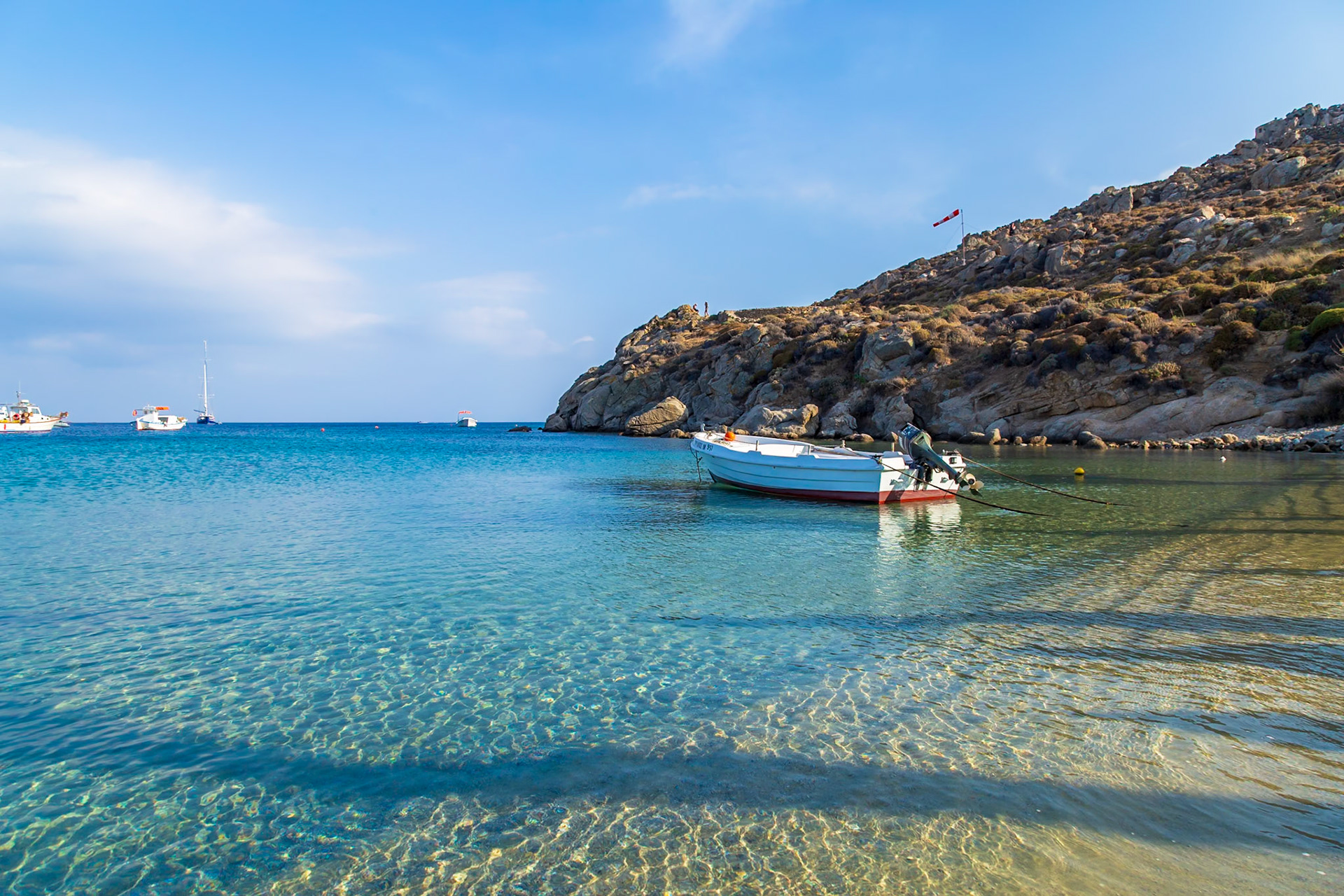Psarou Beach, Mykonos, Greece - May 24th 2018: A small boat is moored in the clear turquoise water, with people on the hill in the background.