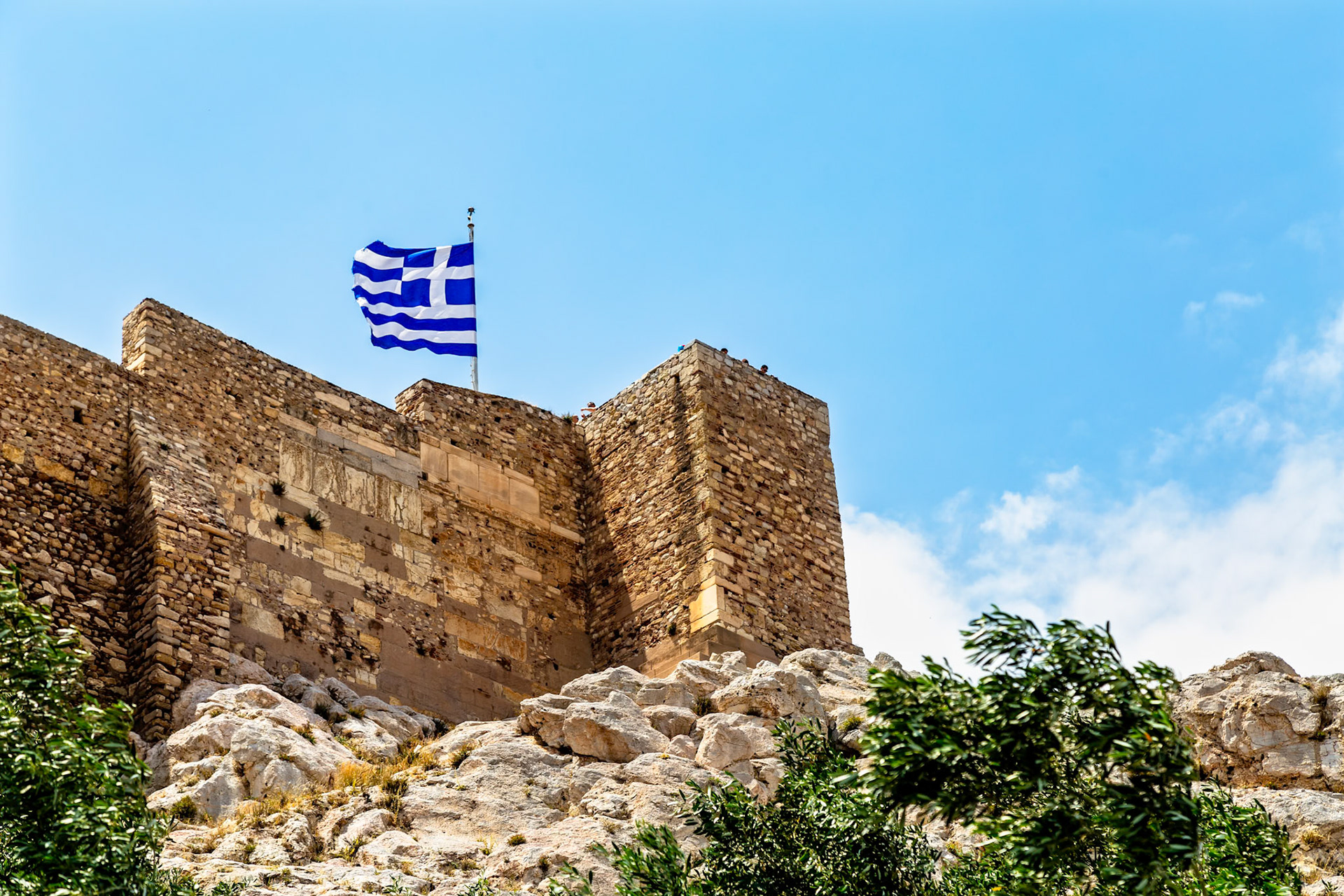 Athens, Greece - May 23rd 2018: The Greek flag flies atop a stone structure, likely part of the Acropolis, symbolizing Greek history and national pride.