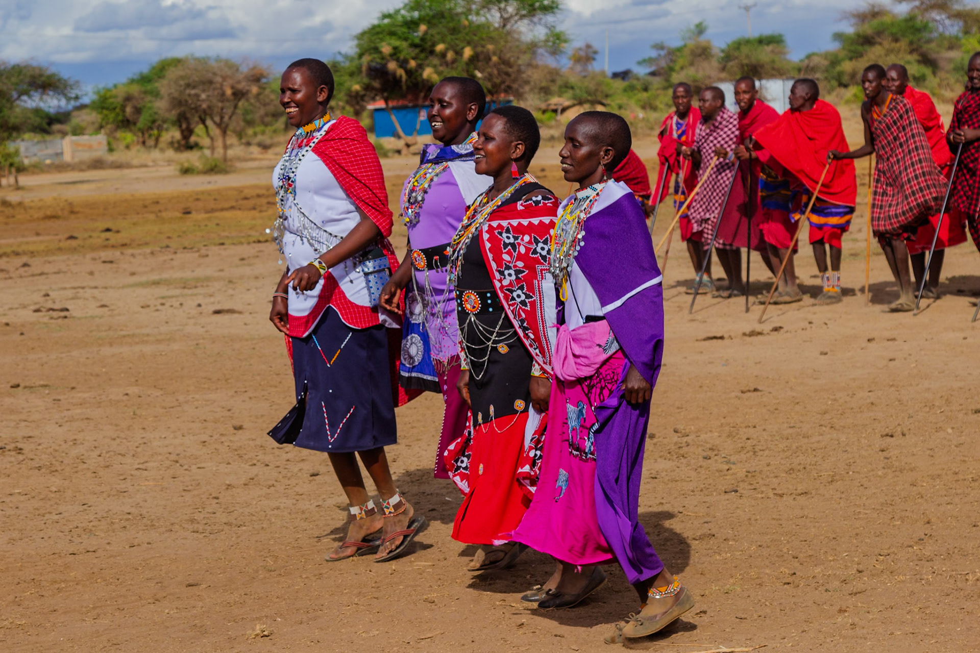 Maasai women in Kenya, adorned in traditional attire, walk with men in a village. The scene captures Maasai culture.