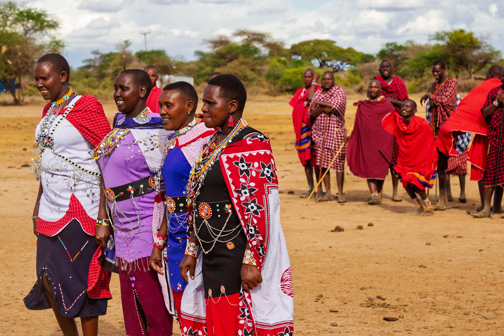 Maasai women in Kenya wear colorful beaded jewelry and patterned cloth while men dance in the background.