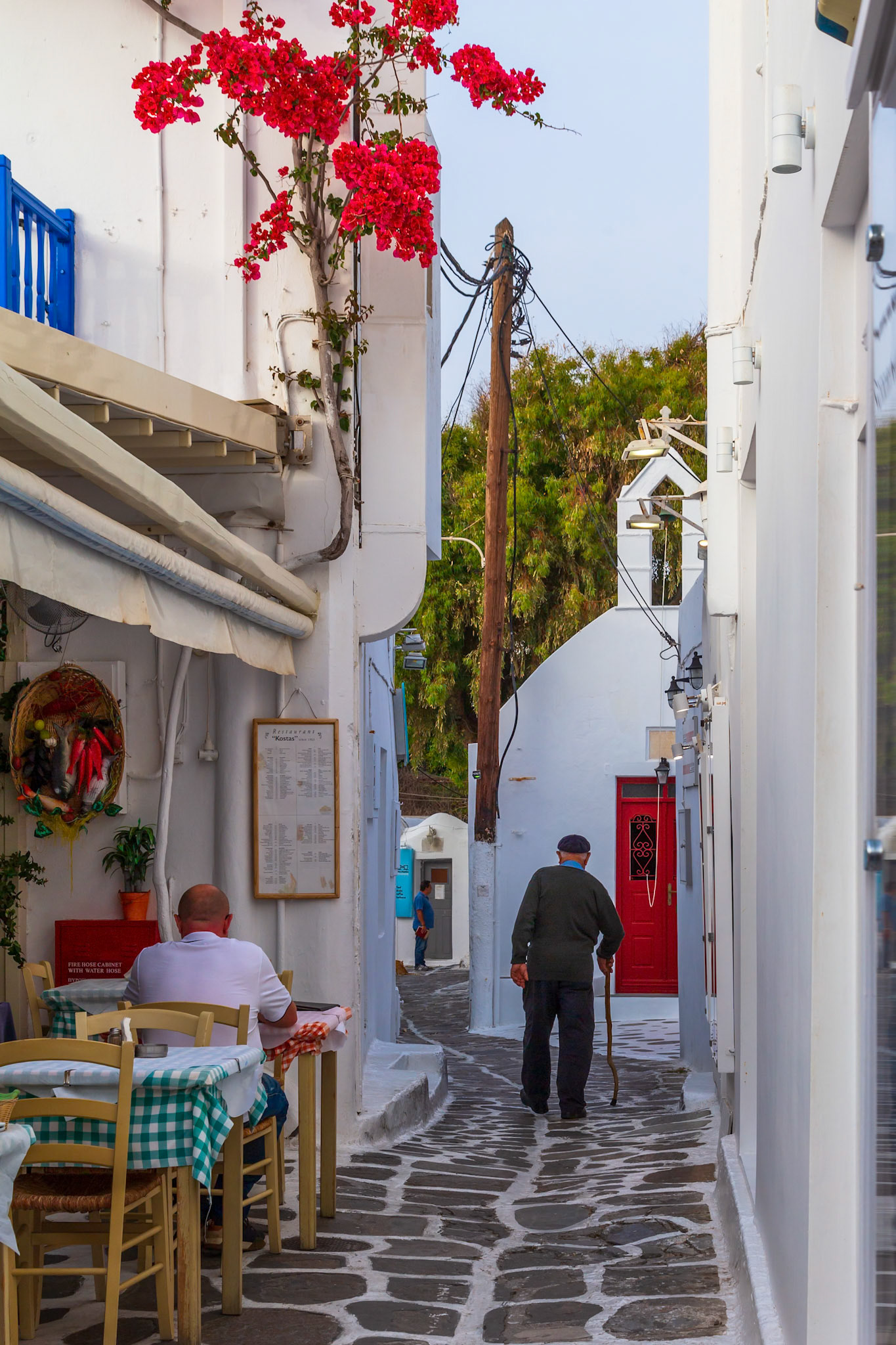 Mykonos, Greece - May 23rd 2018: An elderly man walks down a narrow street, past a restaurant, towards a church with a red door.