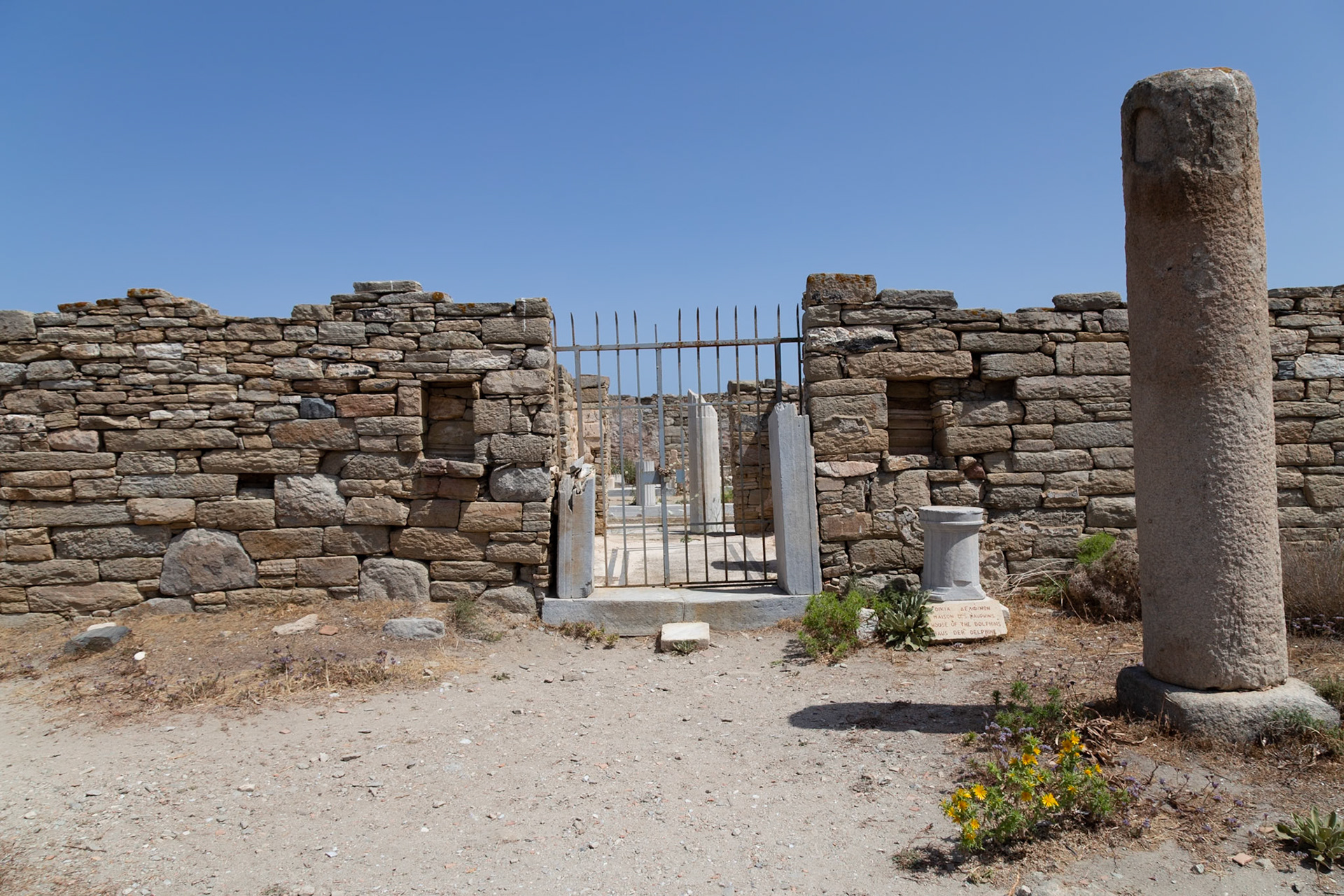 Delos, Greece - May 22nd 2018: A metal gate stands between stone walls, leading to ancient ruins. This is the House of the Dolphins, a historic site.