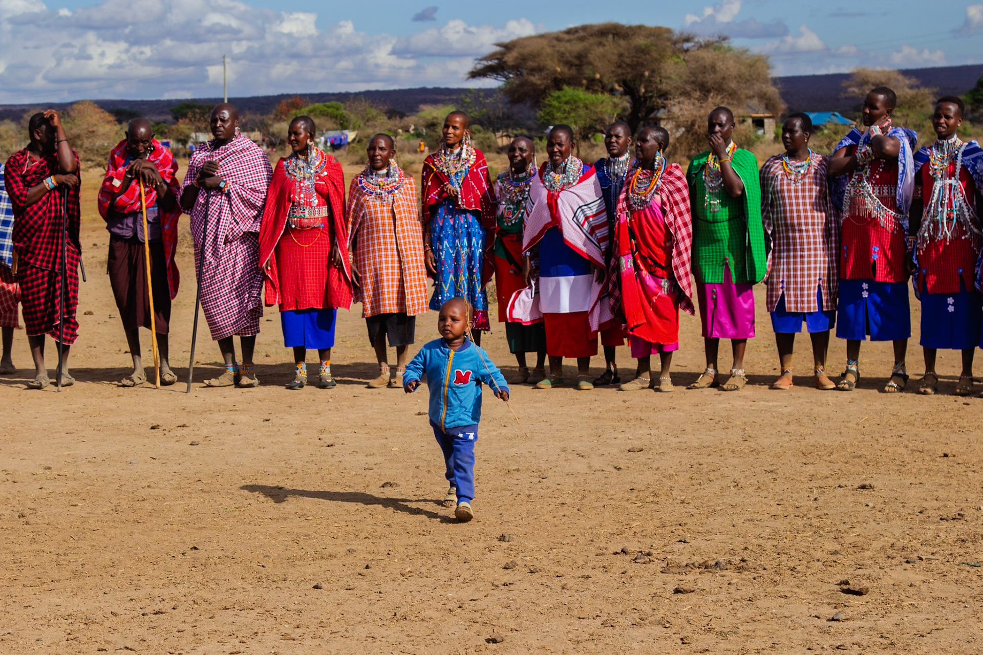 A young child runs in front of Maasai people in traditional clothing in a village in Kenya.