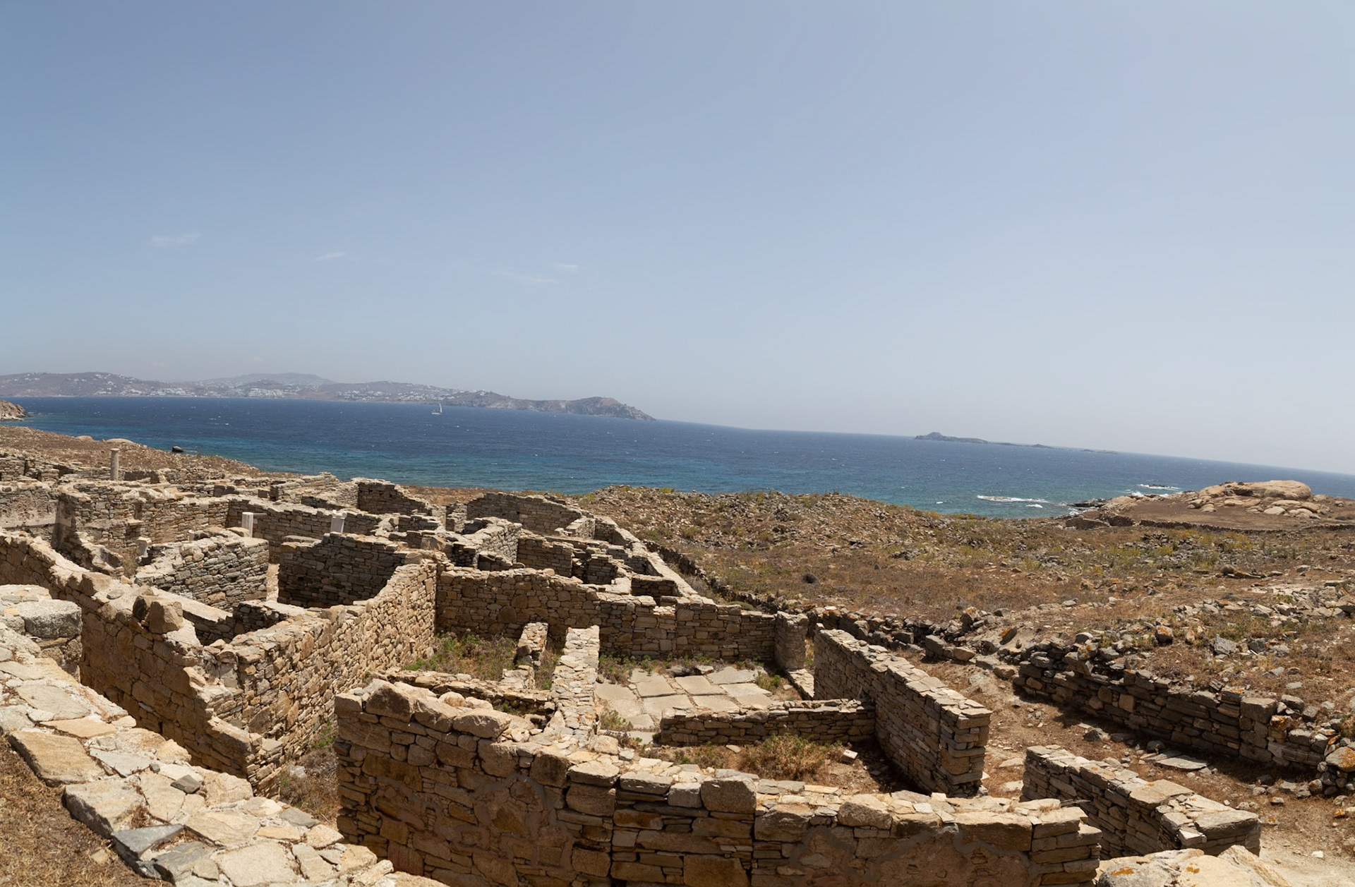 Delos, Greece - May 22nd 2018: Ruins of ancient stone structures stand on Delos, a Greek island, with the Aegean Sea and distant islands visible in the background.