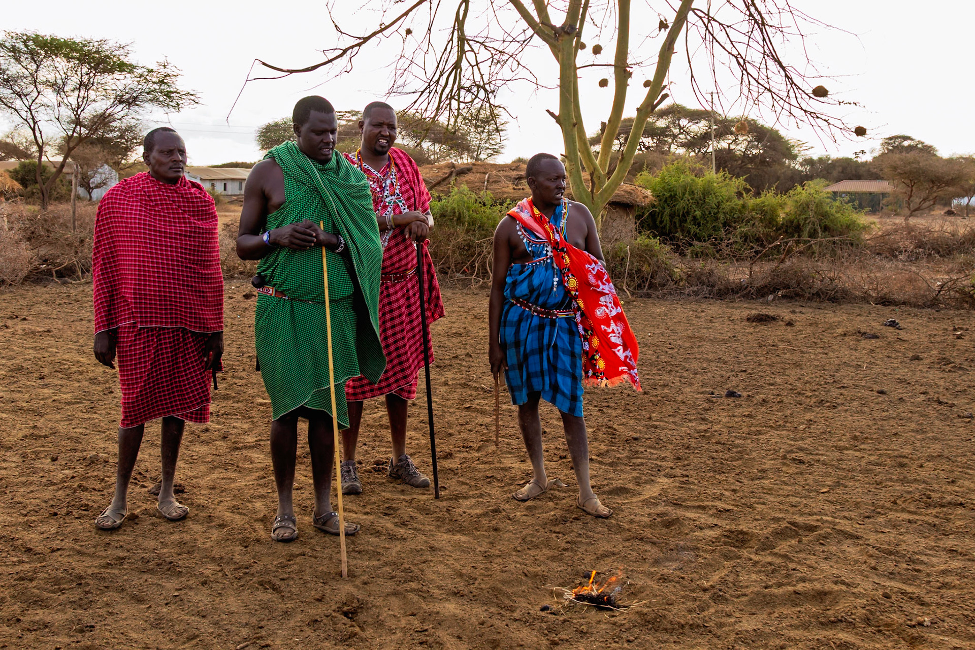 Maasai men in Kenya gather around a small fire, wearing traditional shuka cloths, likely for a ceremony or community meeting.