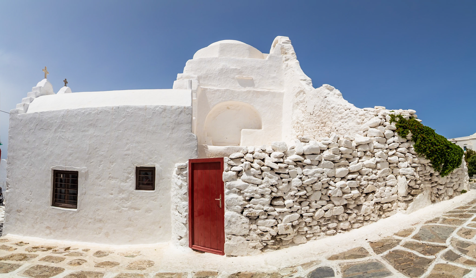 Mykonos, Greece - May 23rd 2018: A traditional whitewashed church with a red door stands against a clear blue sky, showcasing Cycladic architecture.