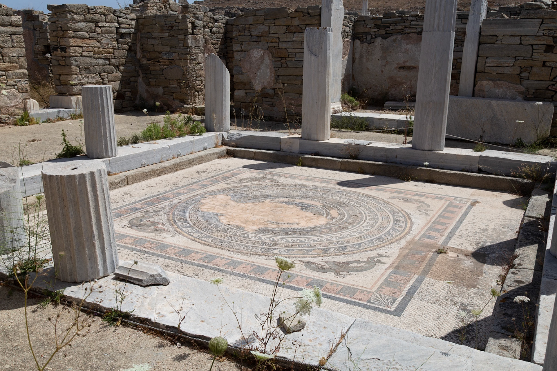 Delos, Greece - May 22nd 2018: A mosaic floor is surrounded by stone walls and columns. The mosaic is decorated with circular patterns and images of sea creatures.