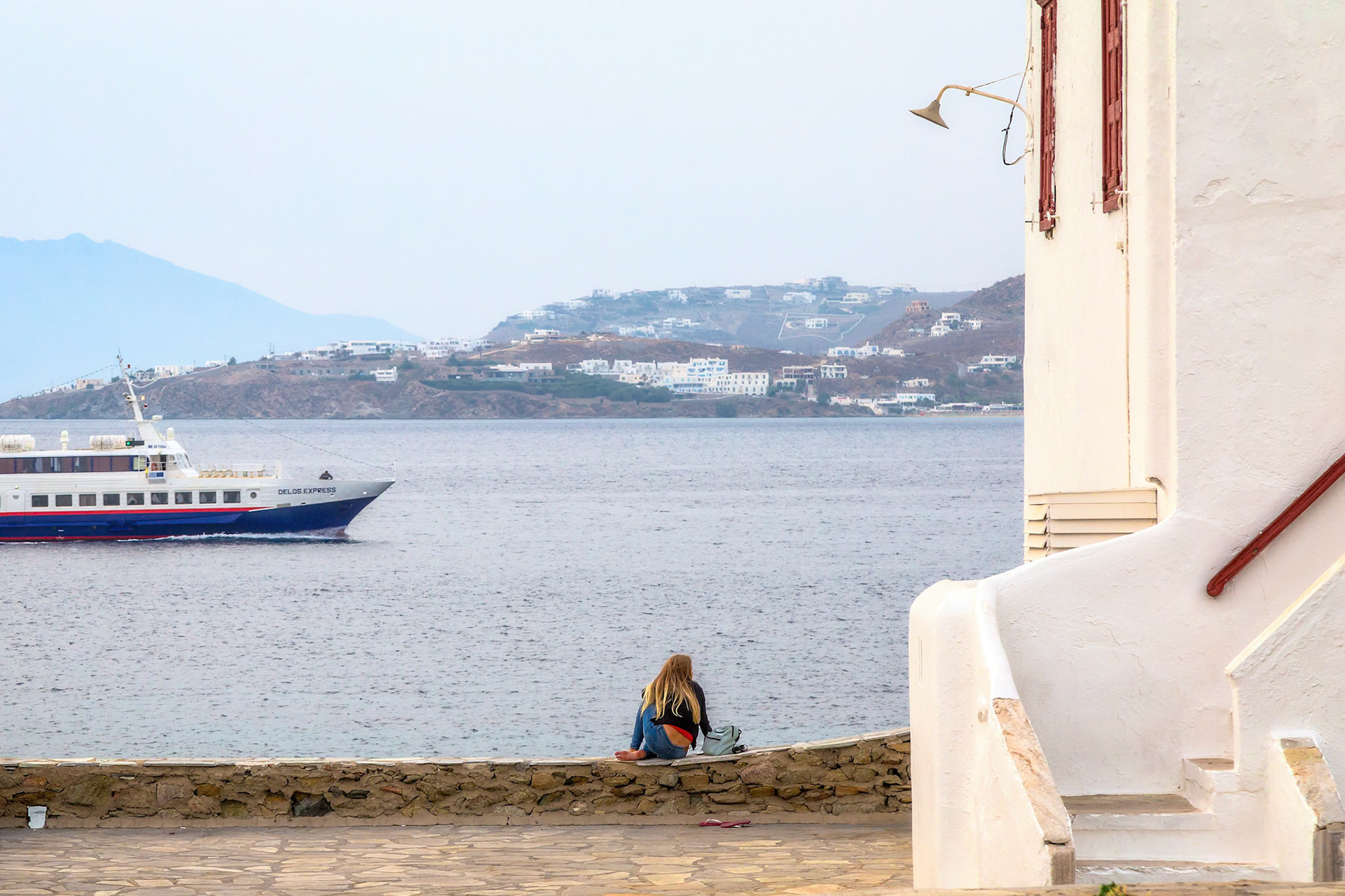 Mykonos, Greece - May 23rd 2018: A woman sits on a wall, watching the Delos Express ferry pass by on the water, enjoying the view of the Greek island.