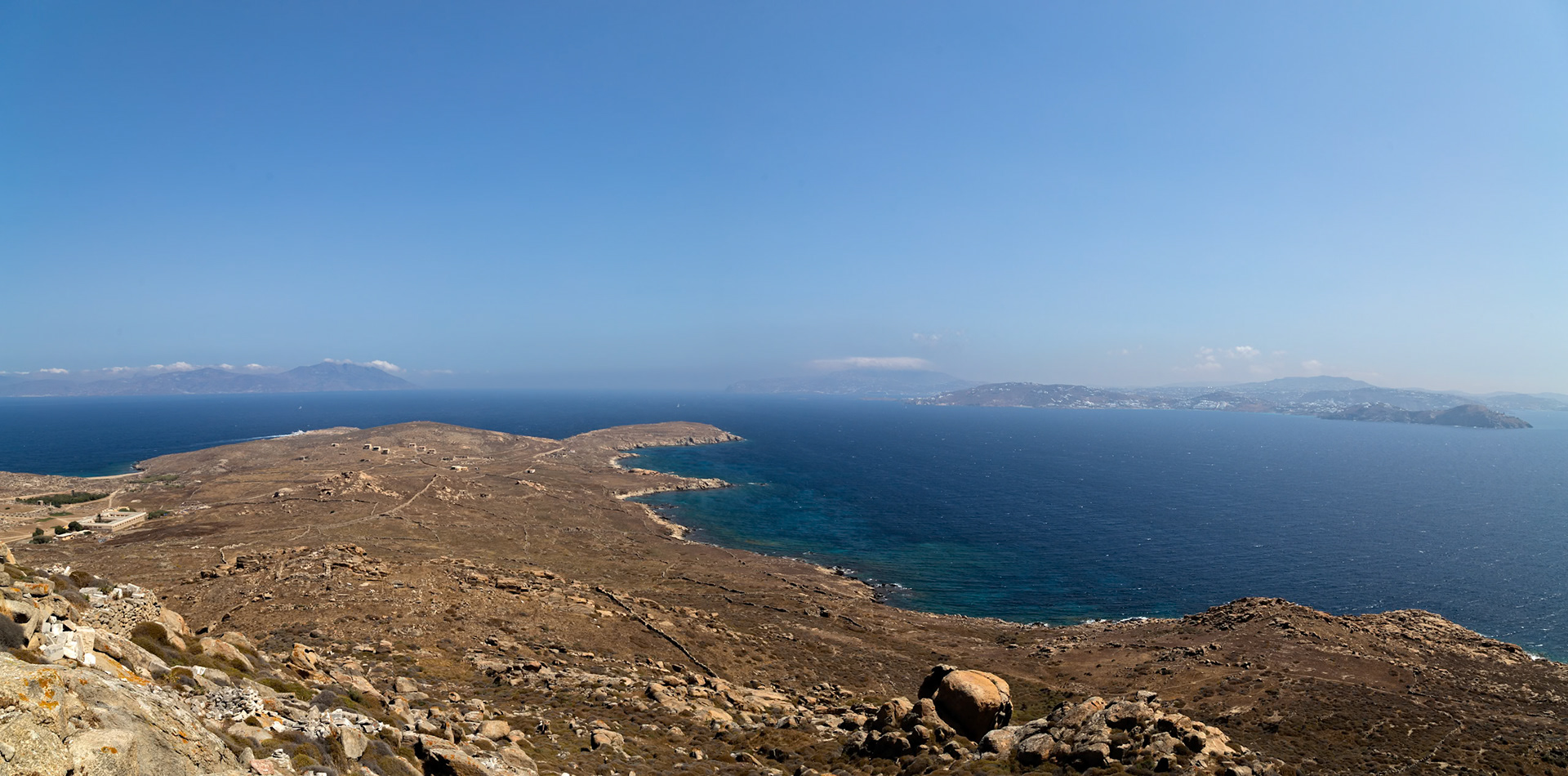 Delos, Greece - May 22nd 2018: A panoramic view of the island of Delos, showcasing its rugged terrain, clear blue waters, and distant islands under a clear sky.