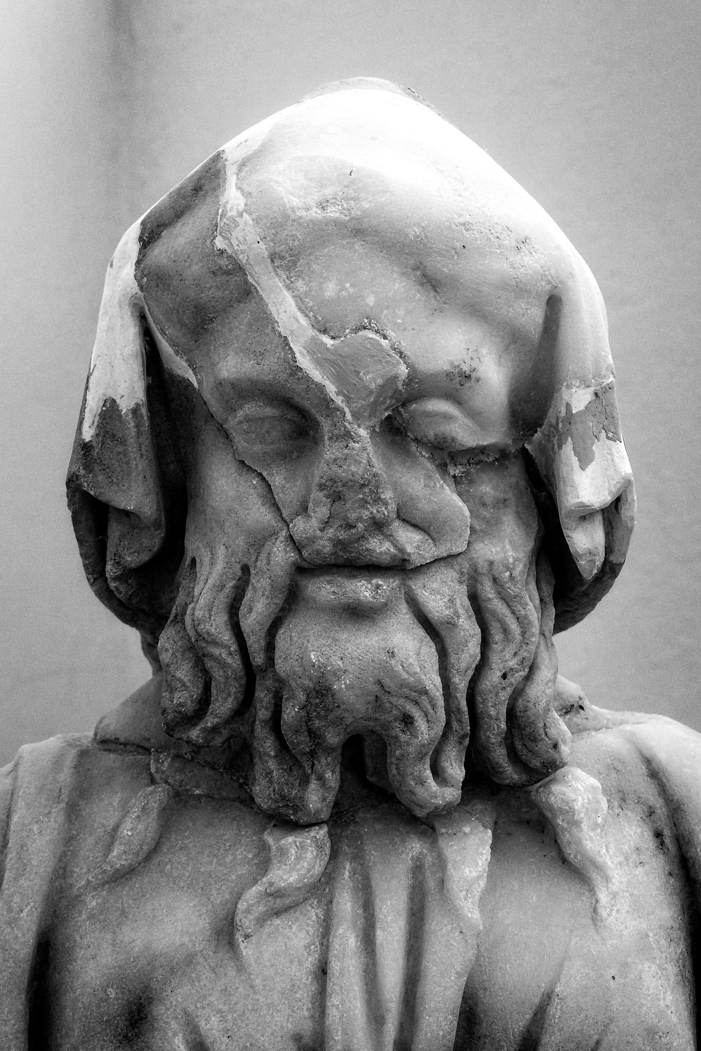 Delos, Greece - May 22nd 2018: A weathered, black and white marble bust of a bearded man with a draped head covering, showcasing ancient Greek sculpture.