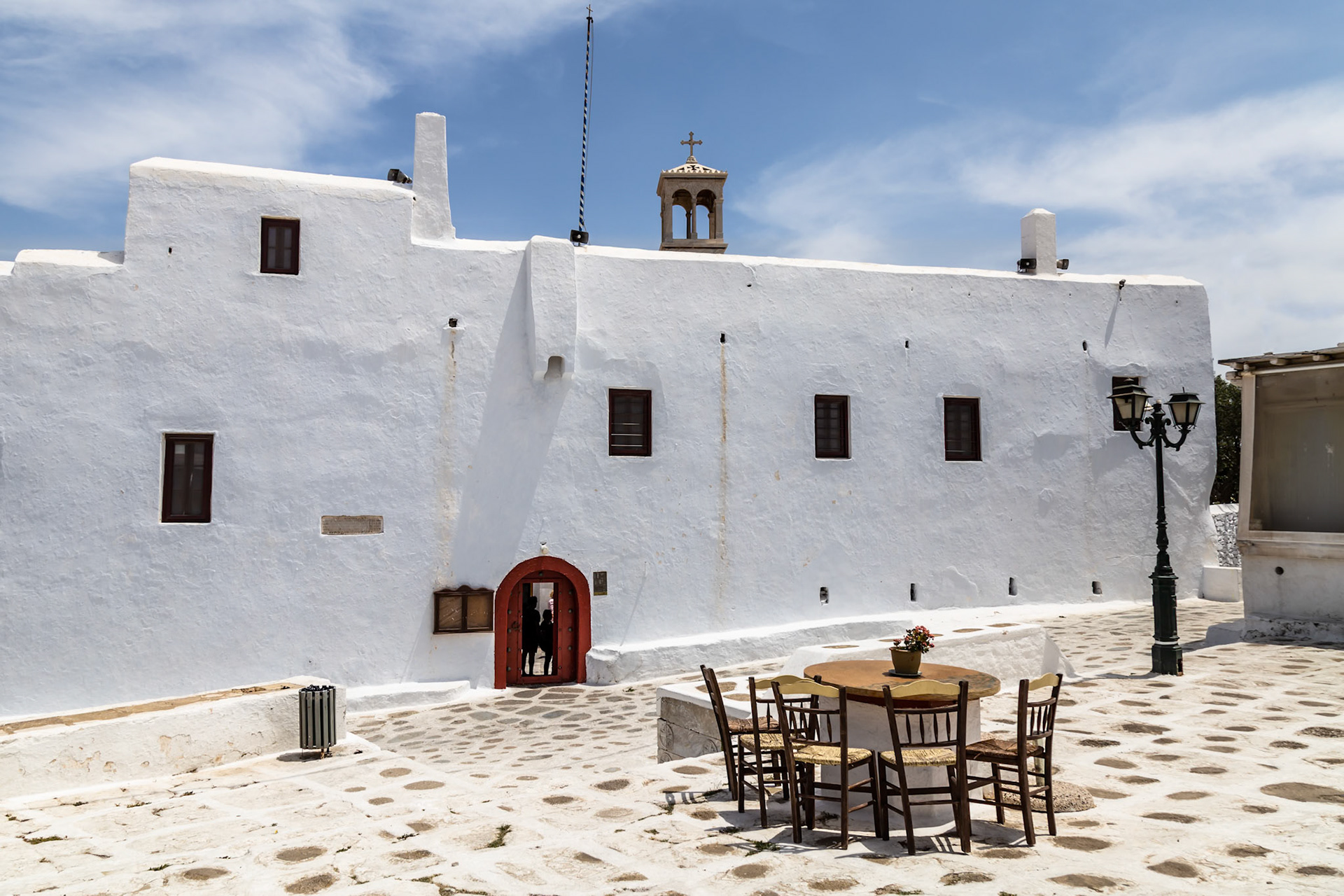 Mykonos, Greece - May 22nd 2018: A traditional whitewashed building with a red door, table and chairs outside, inviting visitors to explore.