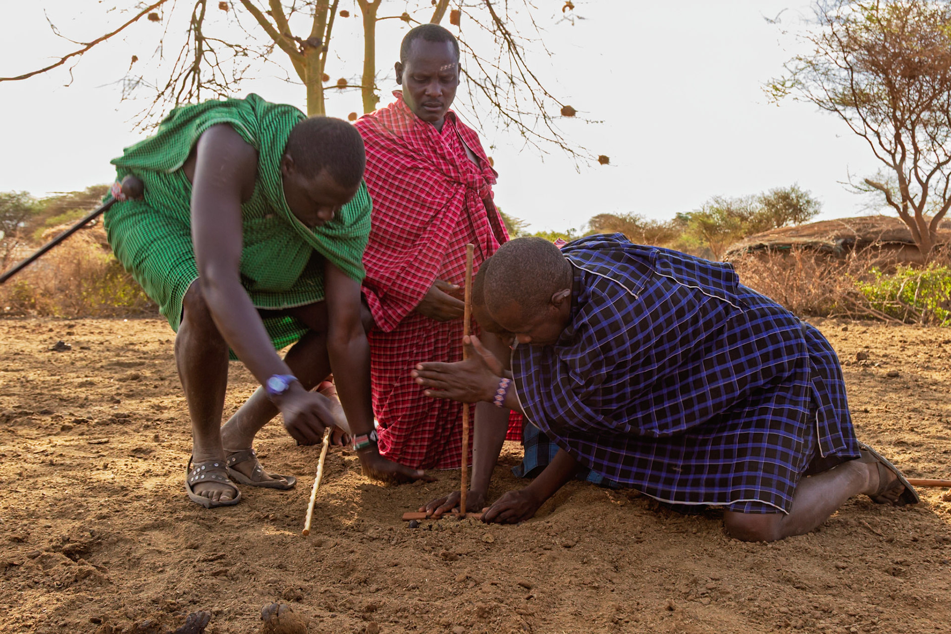 Maasai men in Kenya use sticks to start a fire, demonstrating traditional skills in their village.