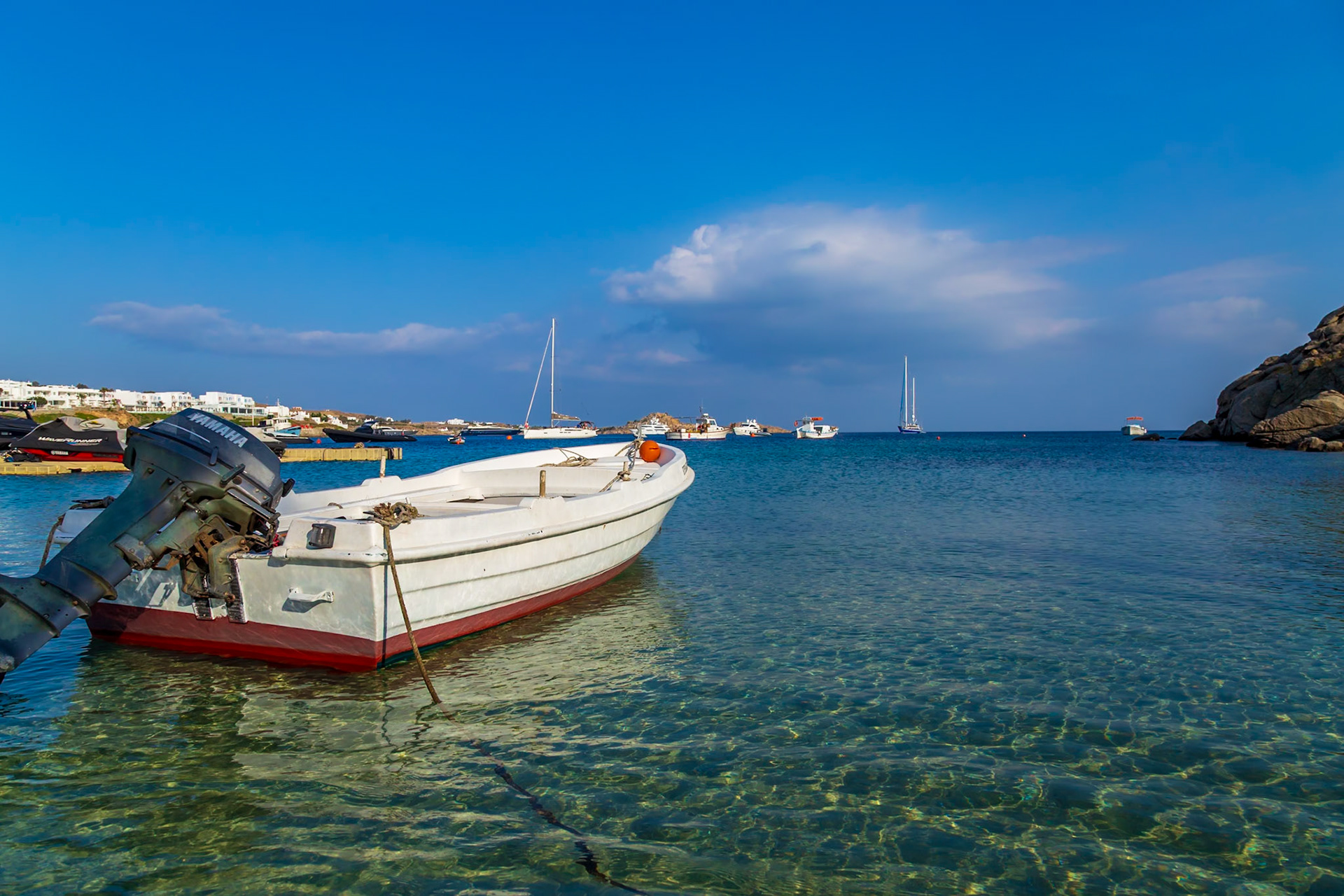 Psarou Beach, Mykonos, Greece - May 24th 2018: A small white boat bobs in the clear turquoise water, other boats are anchored in the distance.