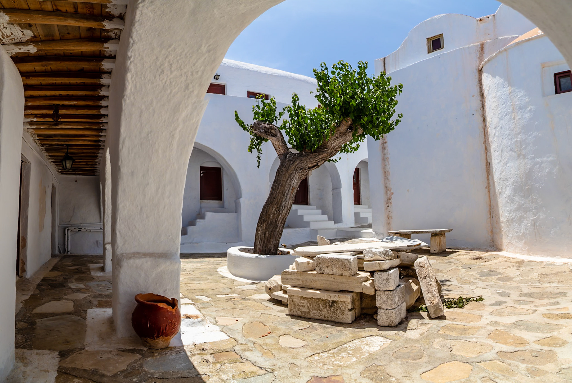 Mykonos, Greece - May 22nd 2018: A courtyard with white buildings, a tree, and stone benches, showcasing traditional Cycladic architecture.