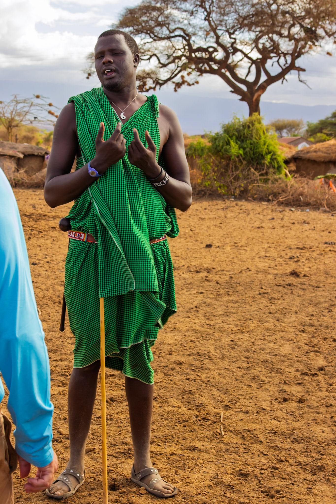 A Maasai man in Kenya, wearing traditional clothing, speaks in his village. He is holding a stick.