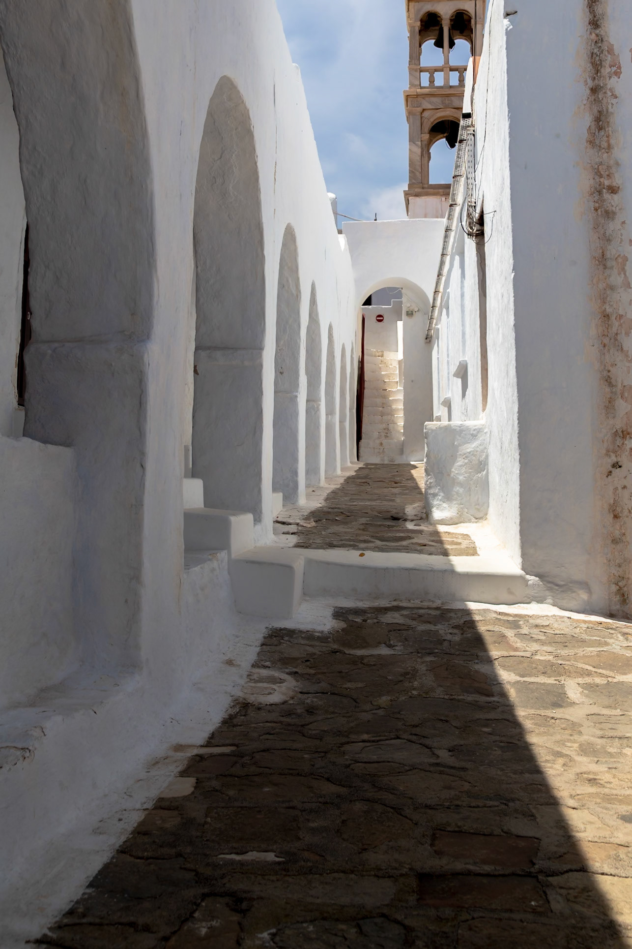 Mykonos, Greece - May 22nd 2018: A narrow, stone-paved alleyway is framed by white-washed buildings, leading towards a distant bell tower.