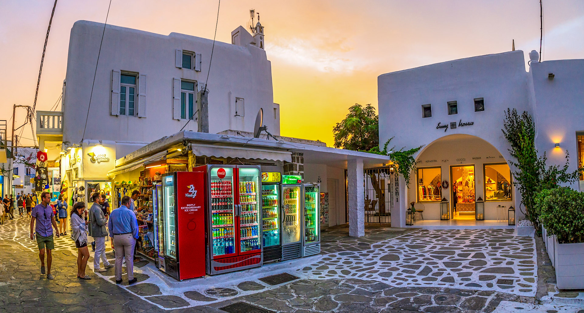Mykonos, Greece - May 22nd 2018: Tourists browse a kiosk selling drinks and ice cream on a street lined with shops at sunset.