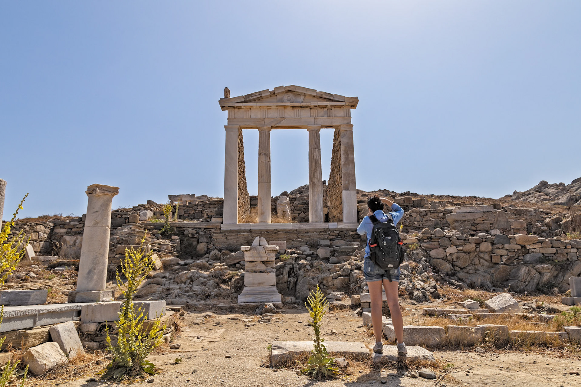 Delos, Greece - May 22nd 2018: A tourist with a backpack is taking a photo of the Temple of Isis ruins. They are visiting the island to explore the ancient archeological site.