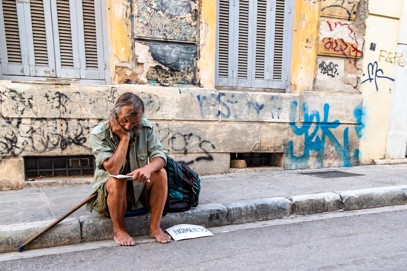 Athens, Greece - May 23rd 2018: A homeless man sits on a curb with a sign, looking down at a piece of paper, possibly contemplating his situation.