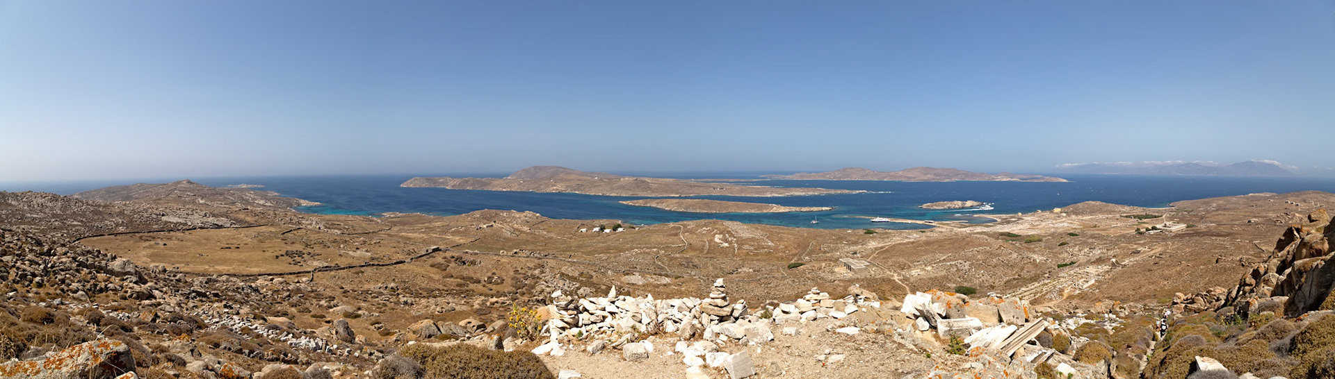 Delos, Greece - May 22nd 2018: A panoramic view of the island of Delos, showcasing its rugged terrain, the Aegean Sea, and neighboring islands under a clear blue sky.