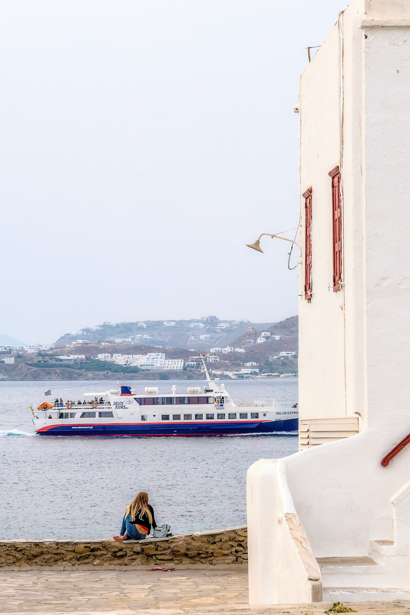 Mykonos, Greece - May 23rd 2018: A woman sits on a wall, watching the Delos Express ferry pass by, enjoying the view and the sea breeze.