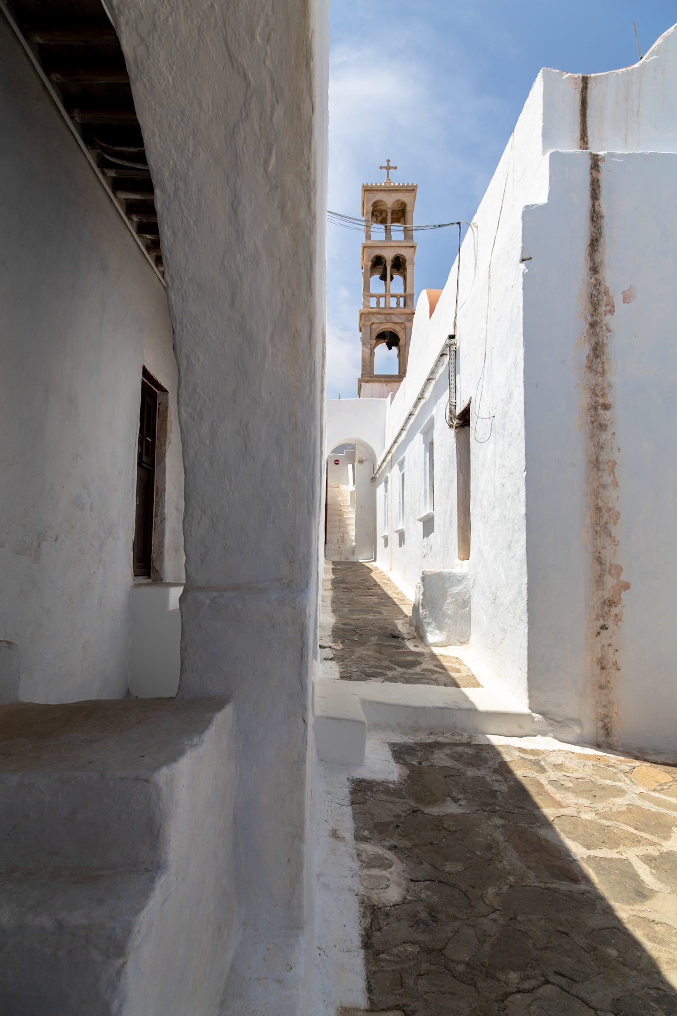 Mykonos, Greece - May 22nd 2018: A narrow alleyway between white buildings leads to a church bell tower, showcasing Cycladic architecture.