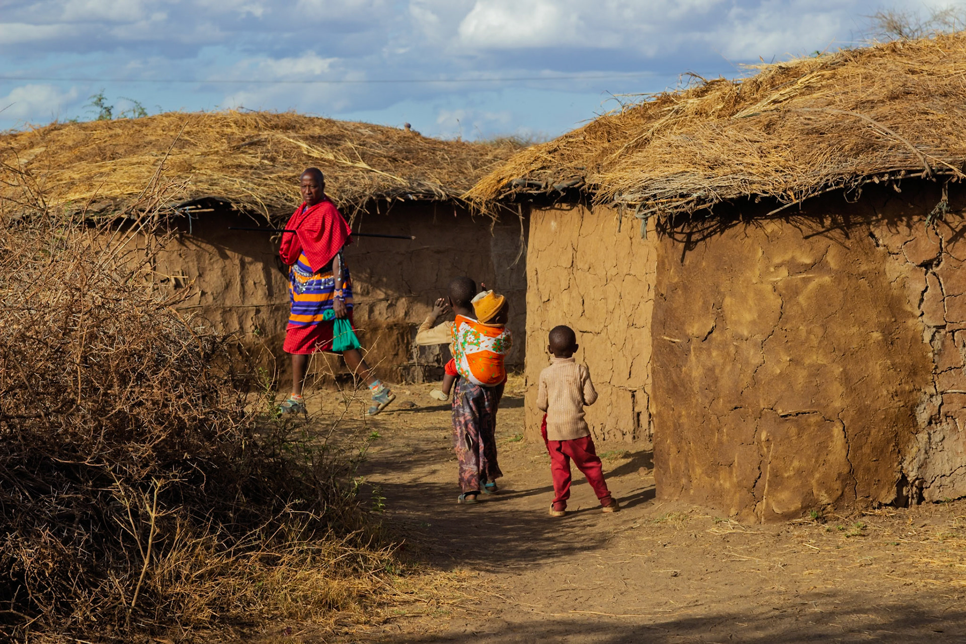 A Maasai village scene in Kenya: A woman carries a child on her back as another child walks nearby, with a Maasai man standing in the background.