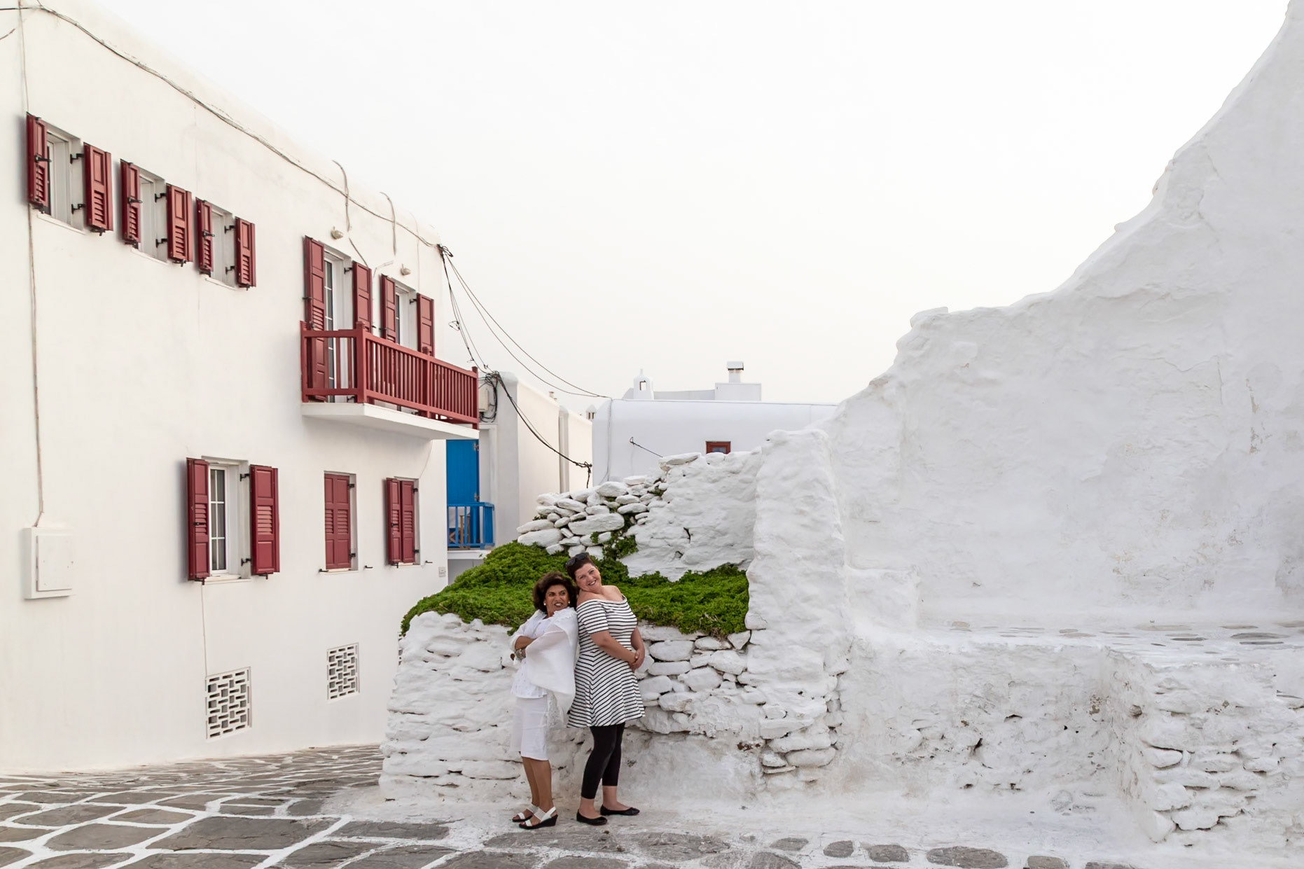Mykonos, Greece - May 23rd 2018: Two women pose for a photo in front of a whitewashed building, embracing and smiling for the camera.