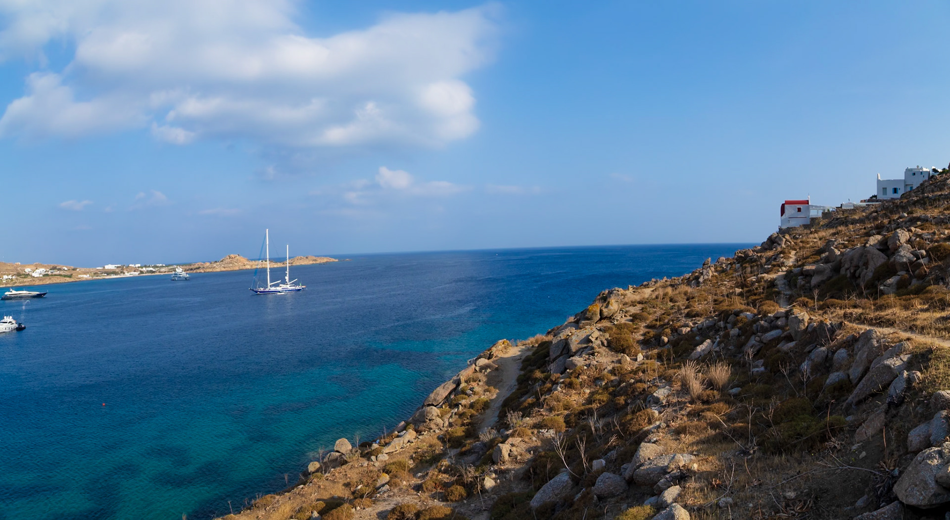 Psarou Beach, Mykonos, Greece - May 24th 2018: Yachts bob in the turquoise waters off the coast, near white buildings on a rocky hillside.
