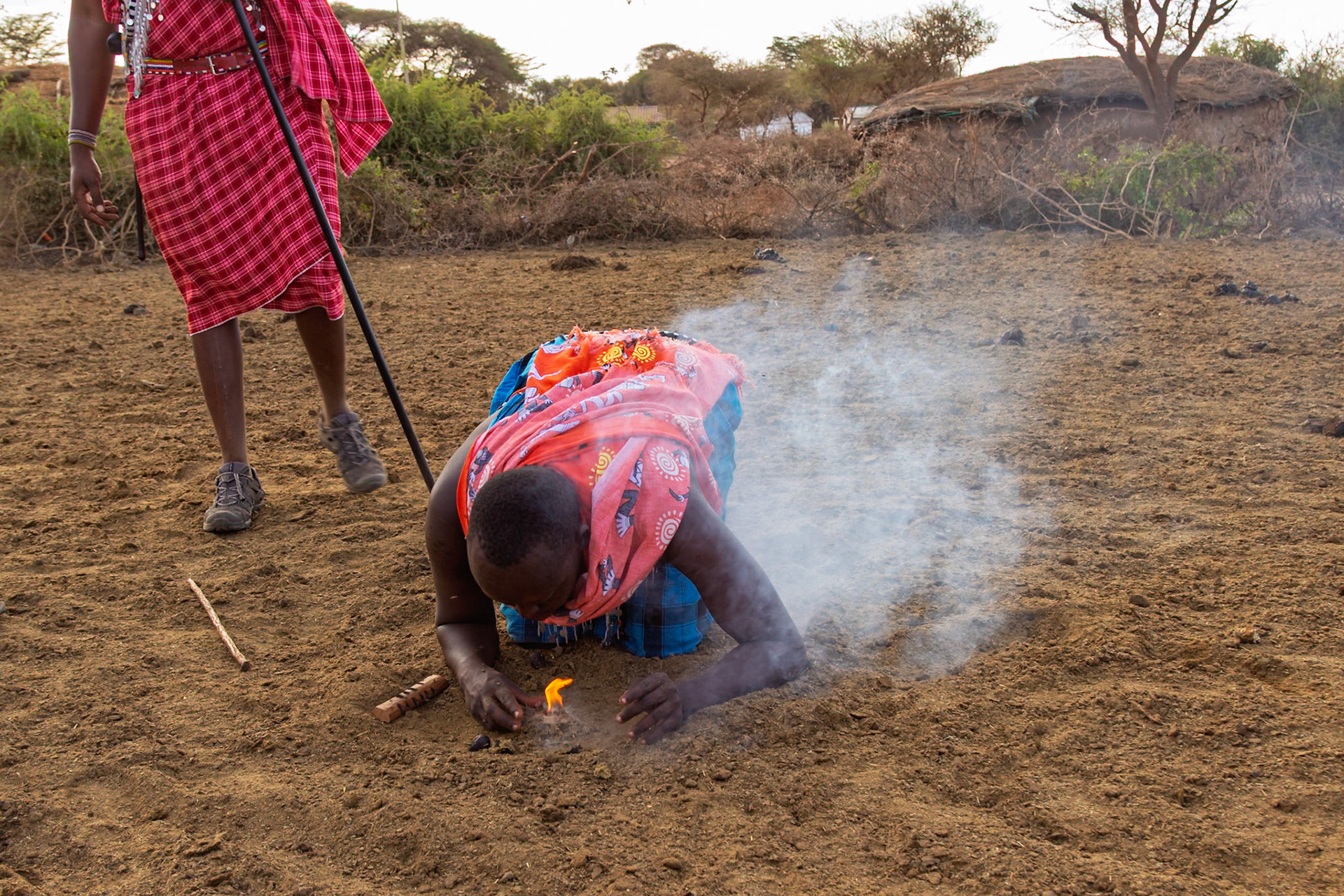 A Maasai man in Kenya starts a fire using traditional methods, while another Maasai man stands nearby.