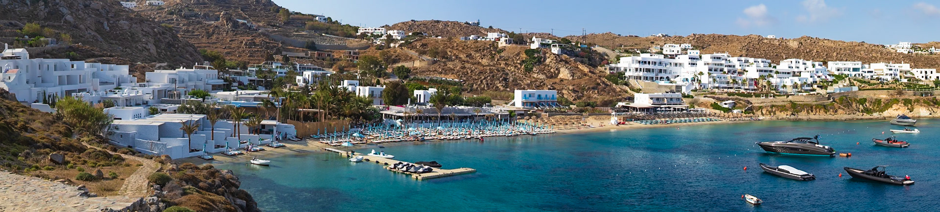 Psarou Beach, Mykonos, Greece - May 24th 2018: A scenic view of the beach, with people relaxing on sunbeds and boats anchored in the turquoise water.