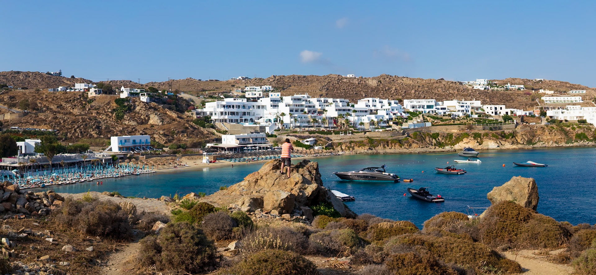 Psarou Beach, Mykonos, Greece - May 24th 2018: A person stands on a rock formation overlooking the beach, boats, and white buildings on the hillside.