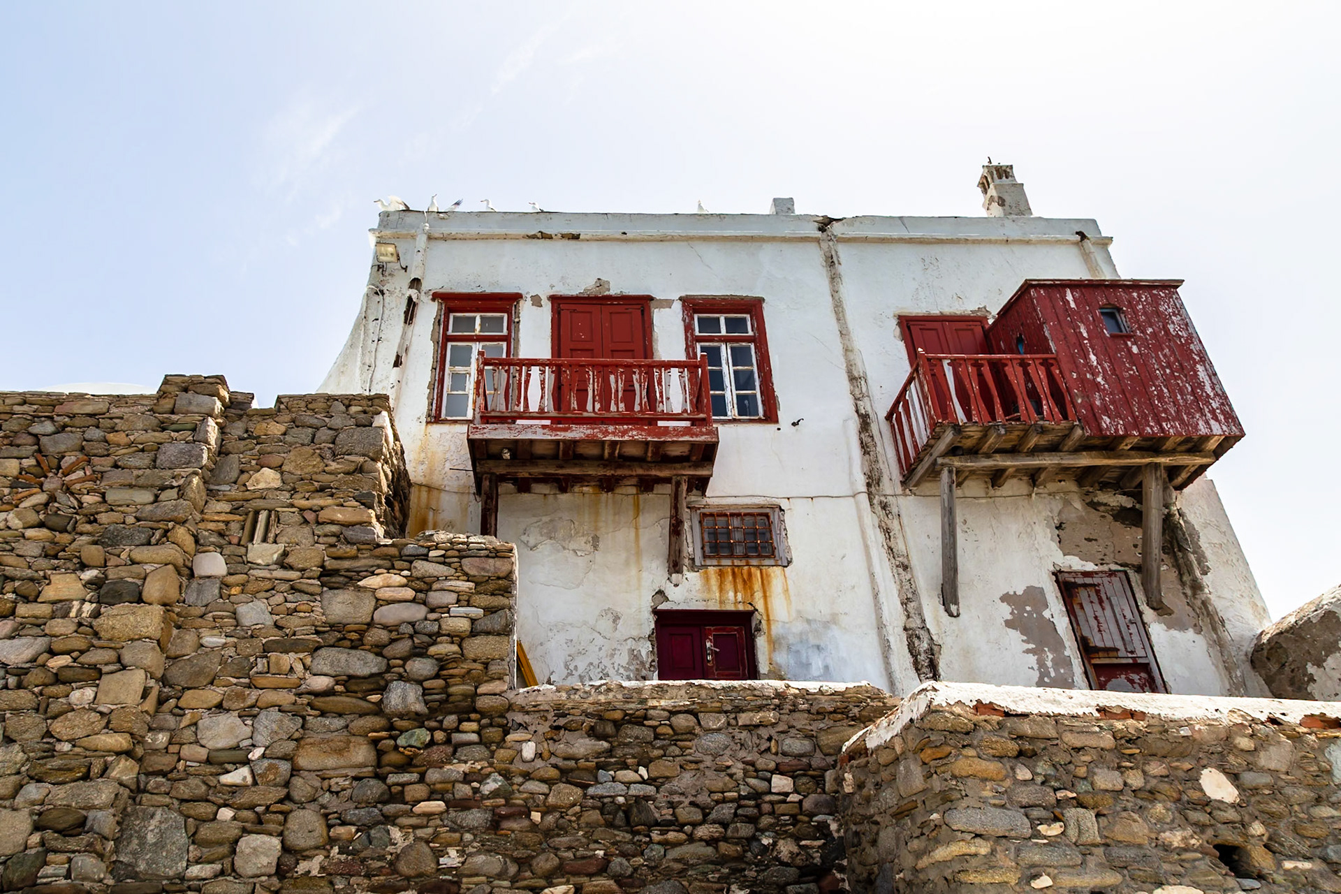 Mykonos, Greece - May 23rd 2018: A weathered building with red balconies and a stone wall shows the unique architecture of the island.
