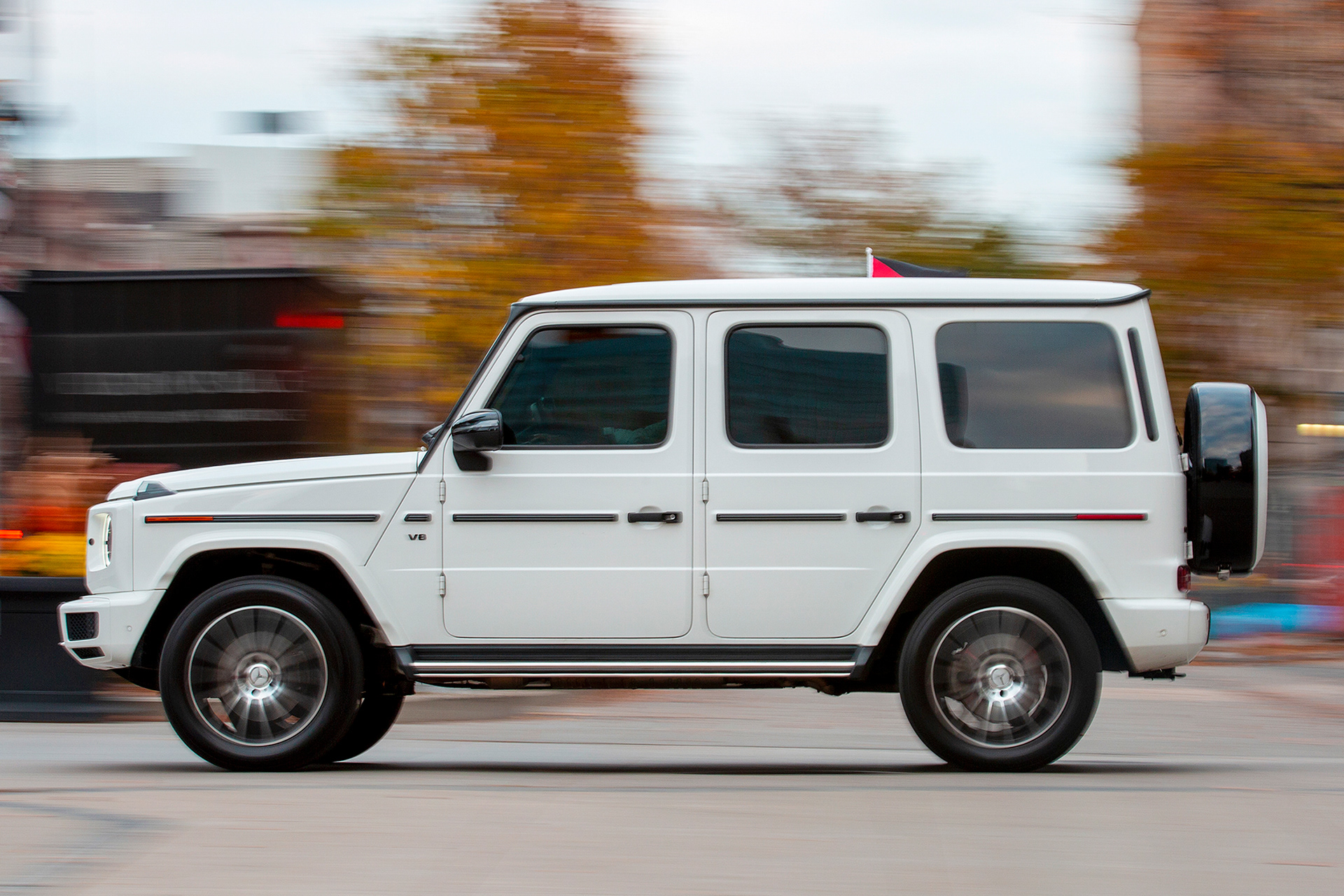 "A white Mercedes-Benz G-Wagon in motion, driving through an urban setting. The background is blurred due to motion blur, emphasizing the vehicle’s movement. The SUV has tinted windows, a spare tire mounted on the rear, and a small flag attached to the roof. Autumn-colored trees and city elements appear in the background."