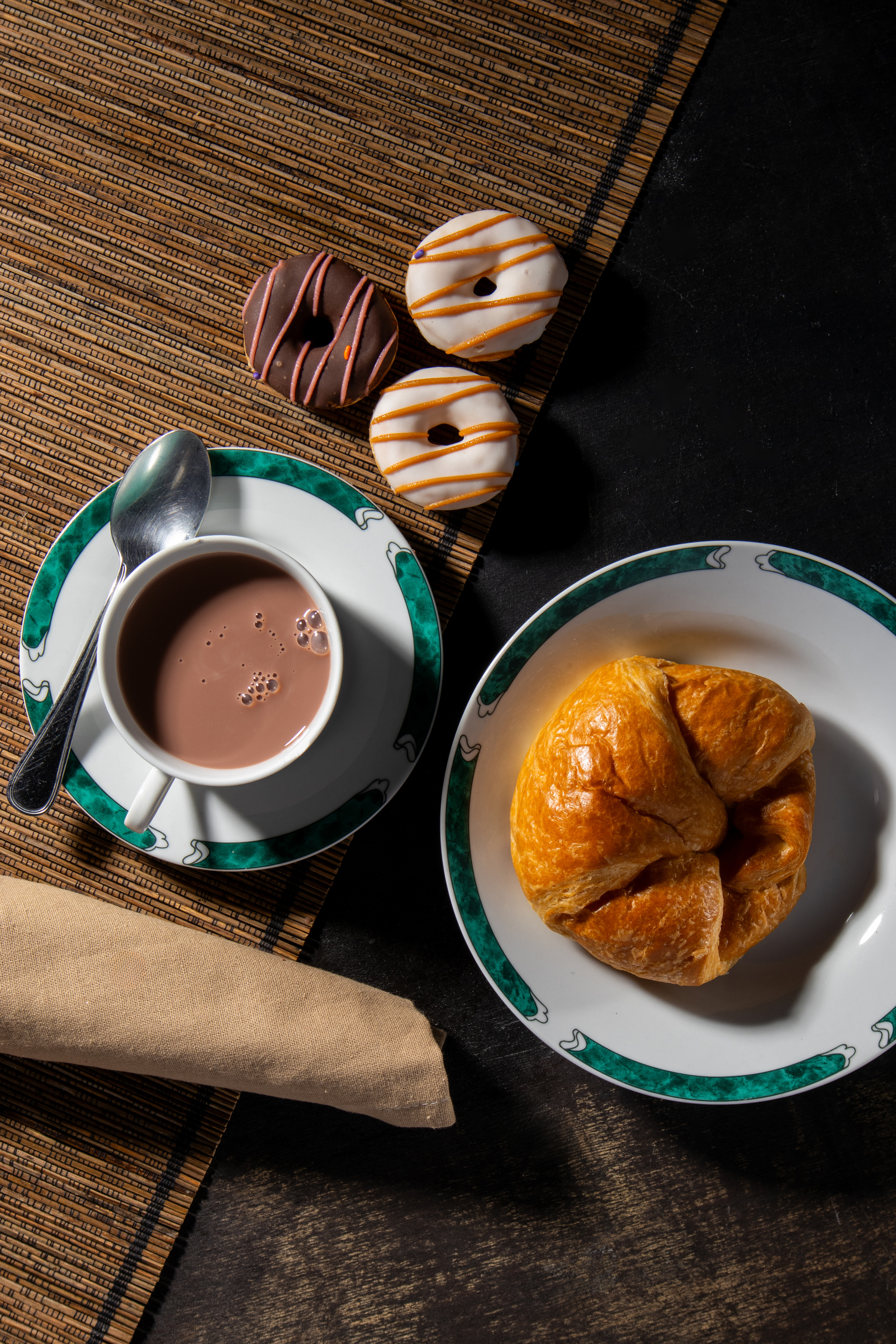"A delicious breakfast spread featuring a golden, flaky croissant on a white plate with a green decorative rim, a steaming cup of hot chocolate with bubbles on the surface, and three small donuts with chocolate and white icing drizzled with colorful stripes. A silver spoon rests on the saucer, and a beige napkin is folded nearby. The meal is set on a textured bamboo placemat over a dark wooden surface, creating a cozy and inviting atmosphere."