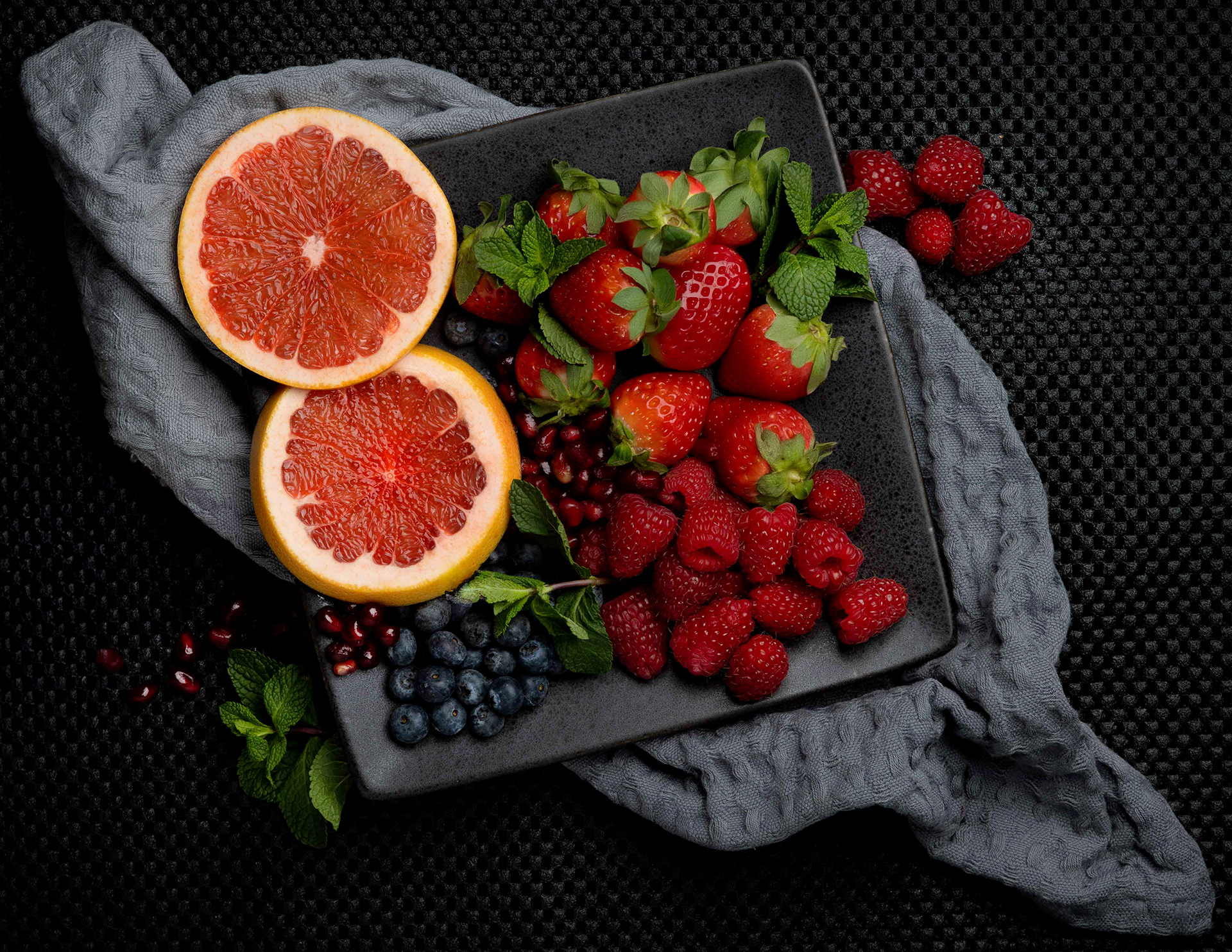 "A beautifully arranged fruit platter on a dark, textured surface, featuring halved grapefruits with a deep red interior, fresh strawberries with green leaves attached, ripe raspberries, blueberries, and pomegranate seeds. The fruits are placed on a black square plate, with a soft gray cloth draped underneath. Fresh mint leaves add a vibrant green contrast to the colorful assortment of fruits."