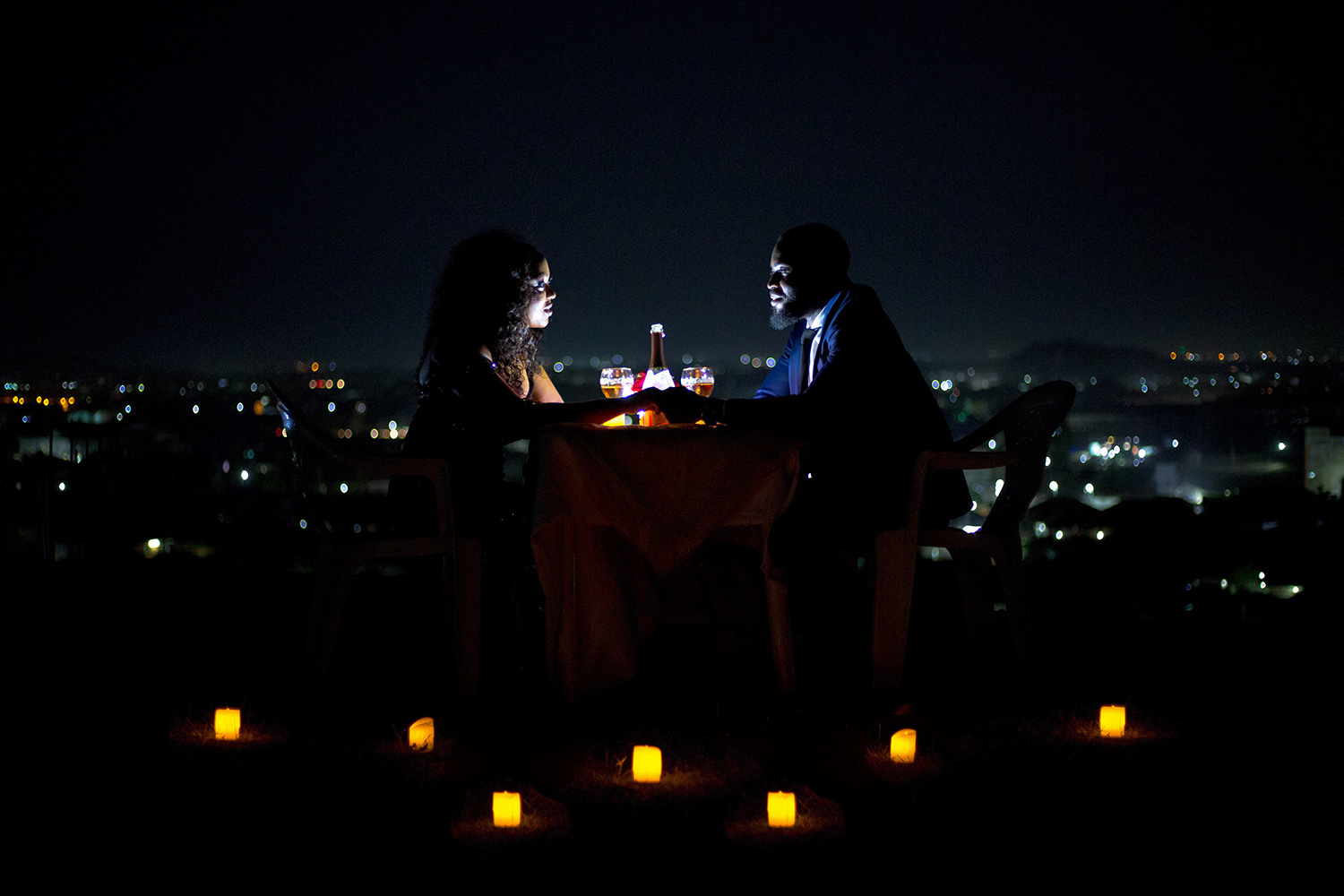 "A couple sits across from each other at a small table on a hilltop at night, sharing a romantic candlelit dinner. They hold hands while gazing into each other's eyes, illuminated by the soft glow of candles scattered around them. A bottle of wine and two glasses sit between them, reflecting the warm light. The background reveals a vast cityscape with twinkling lights, creating a dreamy, intimate atmosphere."