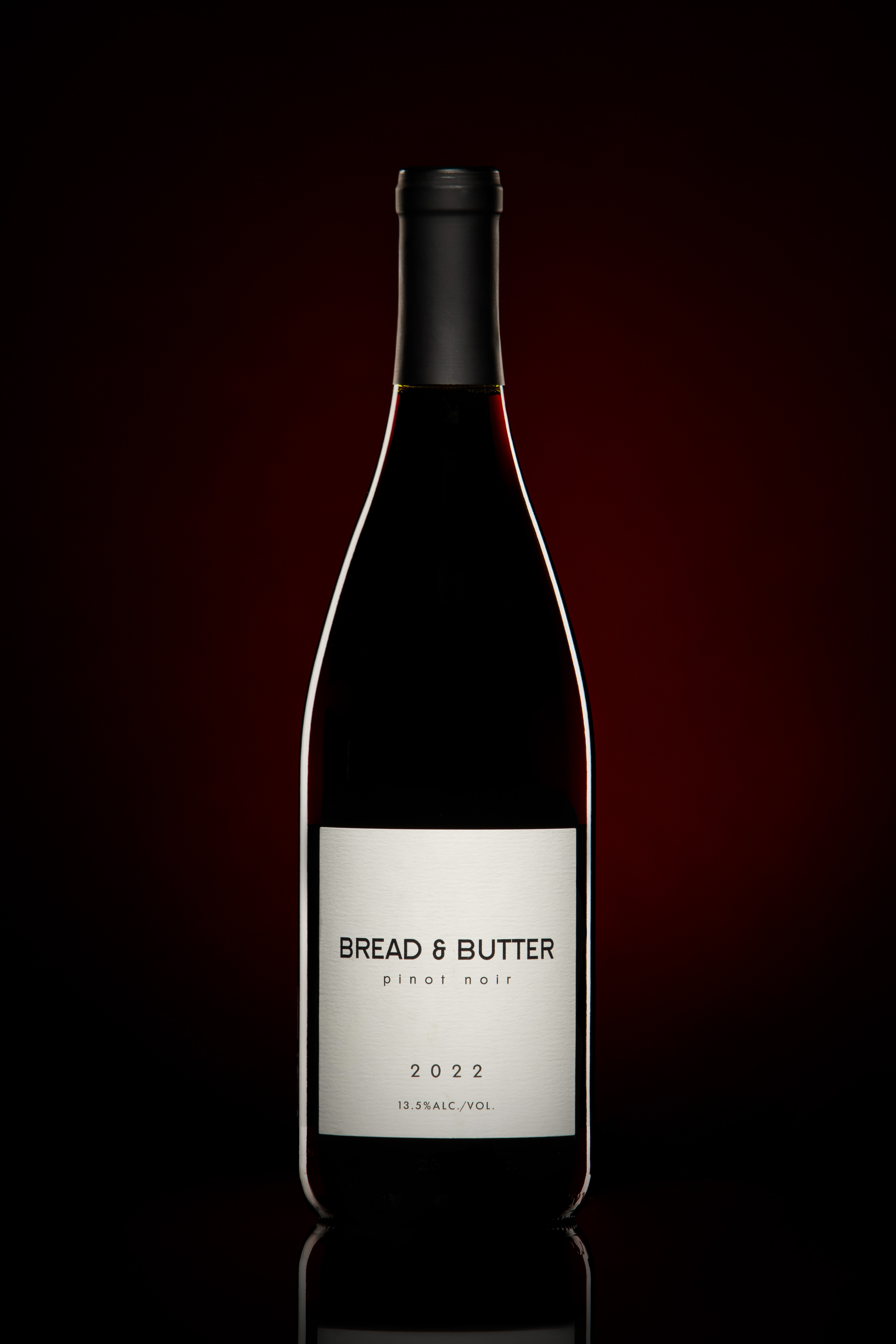 "A bottle of Bread & Butter Pinot Noir 2022 wine, featuring a minimalist white label with black text, set against a dark red gradient background. The label indicates an alcohol content of 13.5%."