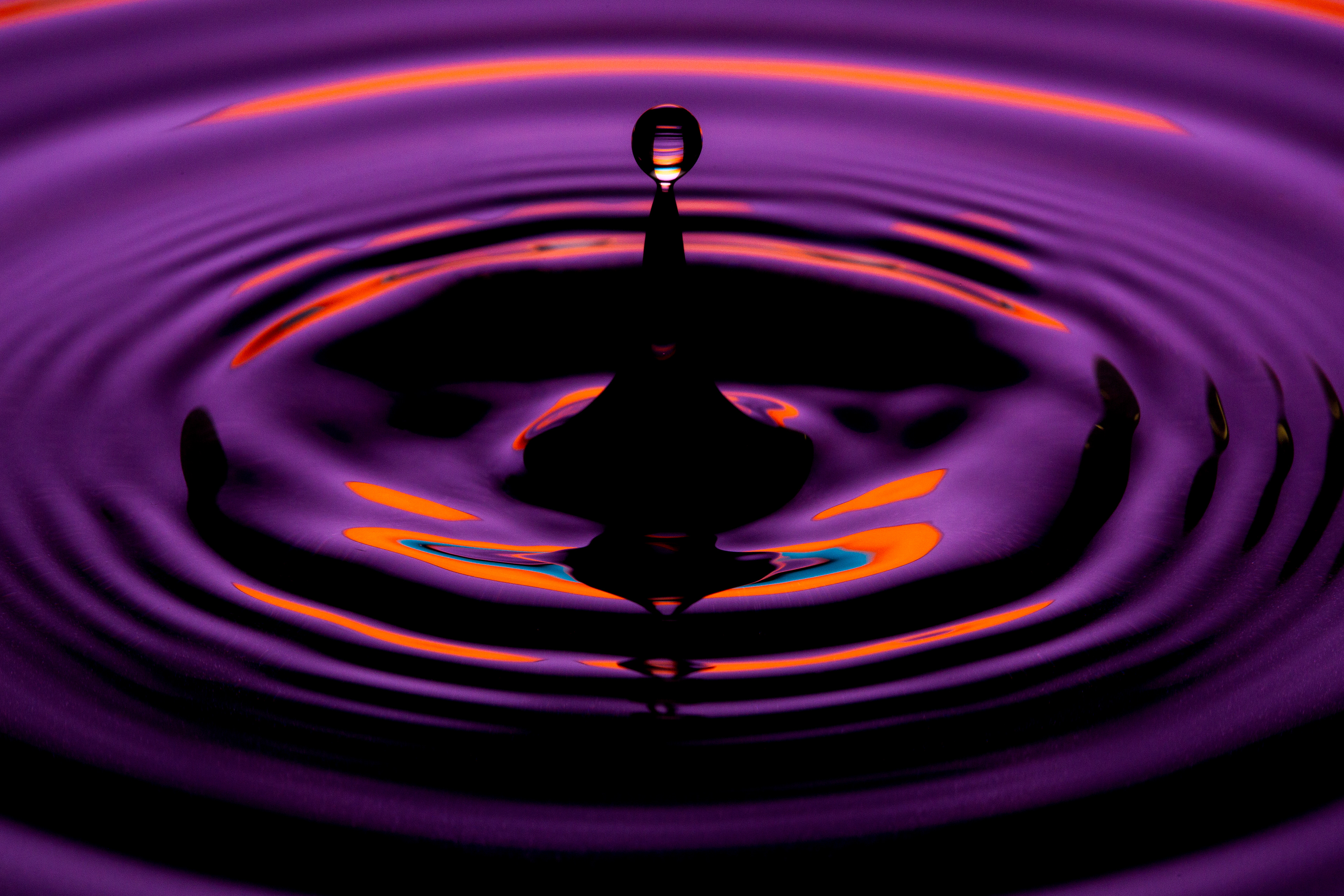 "A striking macro photograph of a water droplet suspended in mid-air just above a conical water splash. The scene is illuminated with vivid purple, orange, and blue reflections, creating a mesmerizing abstract effect. The ripples radiate outward in smooth, concentric waves, enhancing the visual impact of the droplet’s motion."