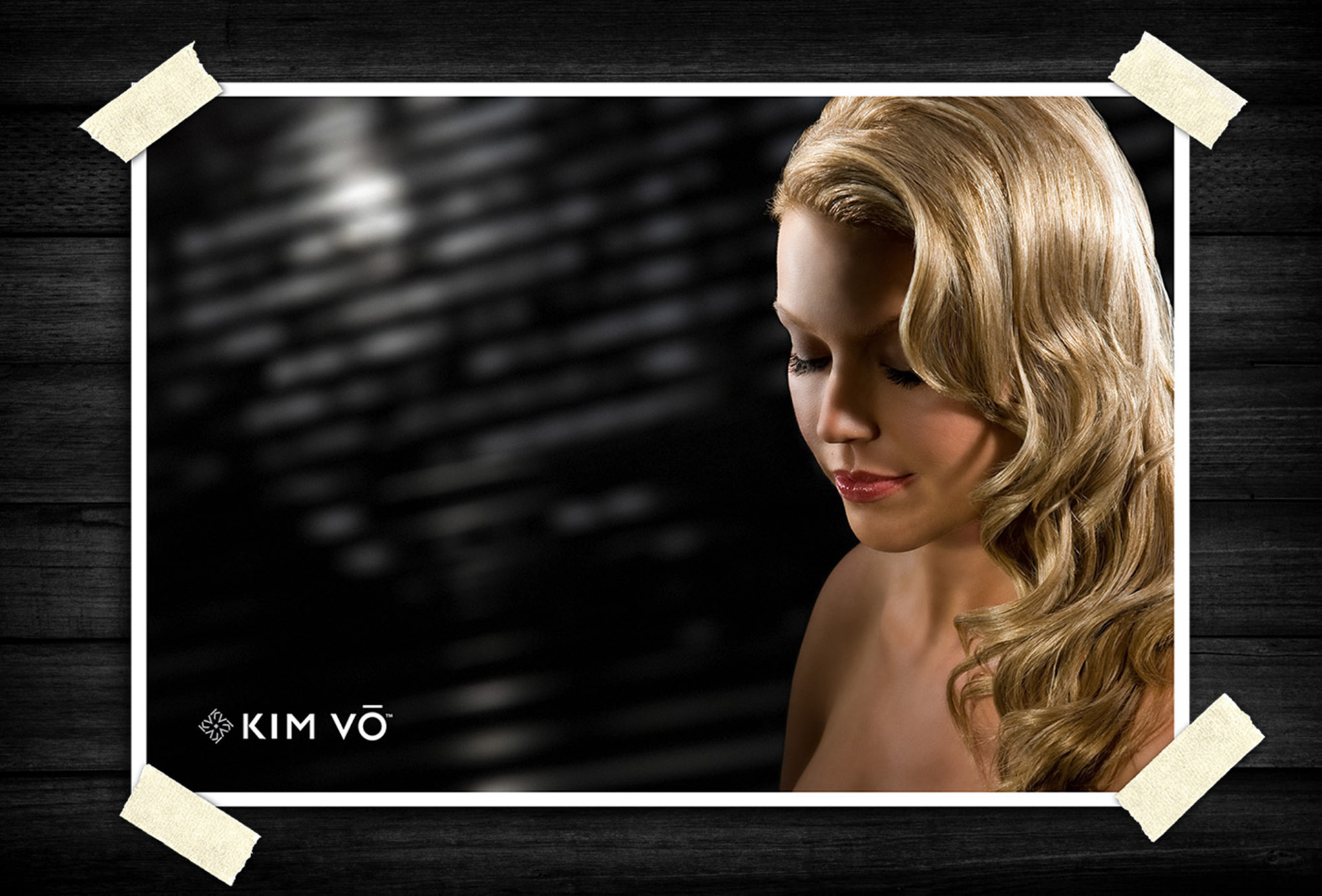 Close-up beauty portrait of a blonde woman with softly curled hair and natural makeup, photographed with dramatic lighting against a dark background.