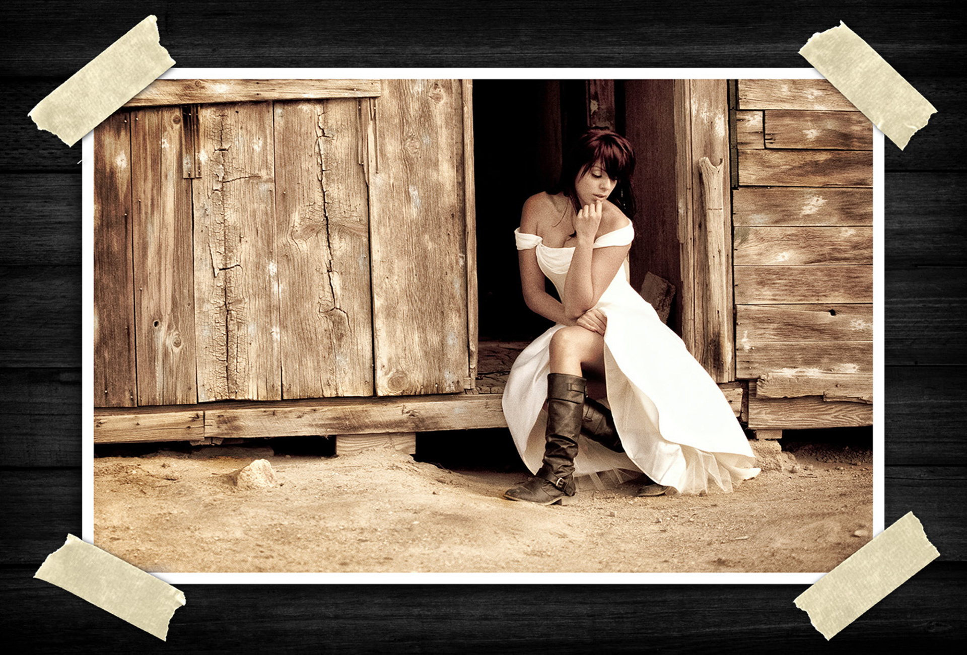 Sepia-toned portrait of a woman in a flowing white dress and tall boots sitting thoughtfully in the doorway of a rustic wooden structure.