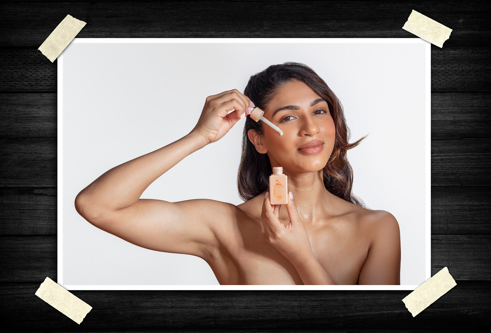 Clean beauty product portrait of a woman applying facial serum with a dropper while holding a skincare bottle against a light studio background.