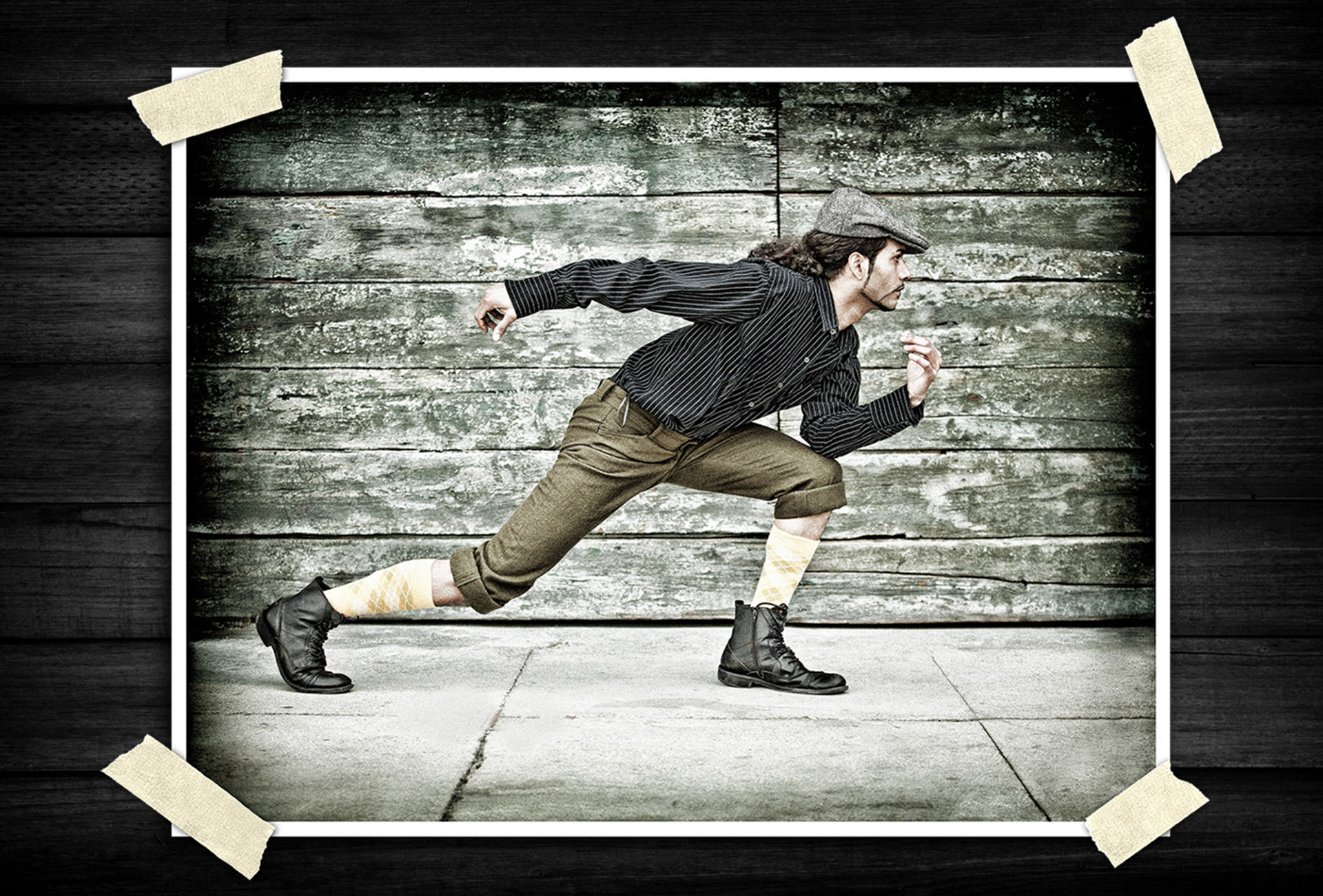 Vintage-inspired portrait of a person in a flat cap and rolled trousers posed mid-stride in a running motion against a weathered wooden wall background.