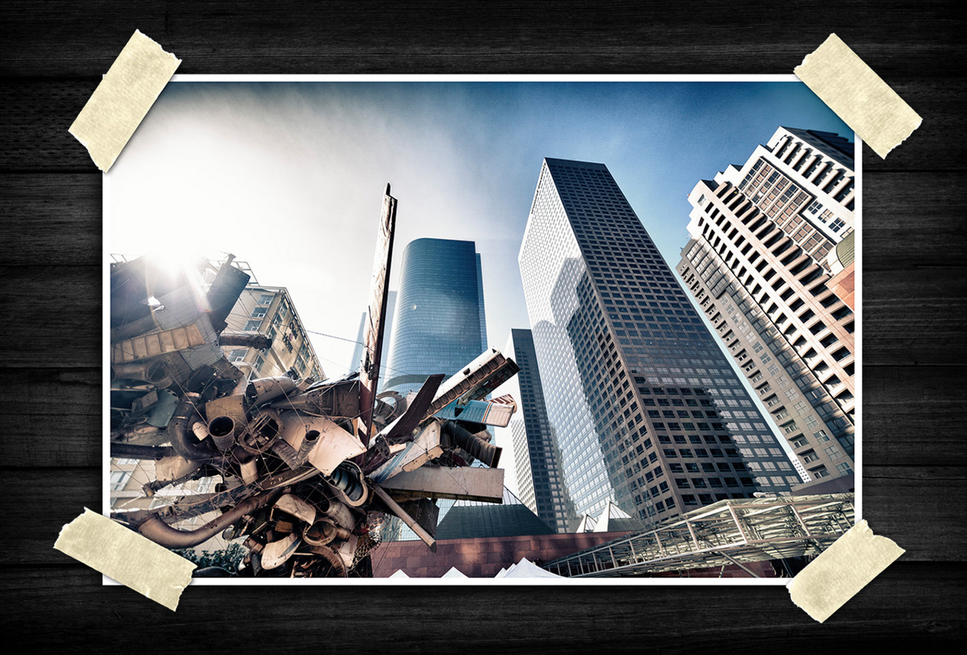 A dramatic cityscape rises from a low-angle perspective, with towering glass skyscrapers stretching into a bright blue sky. An abstract metal sculpture fills the foreground with layered industrial shapes and textures, while strong sunlight creates bold highlights and reflections across the urban scene.
