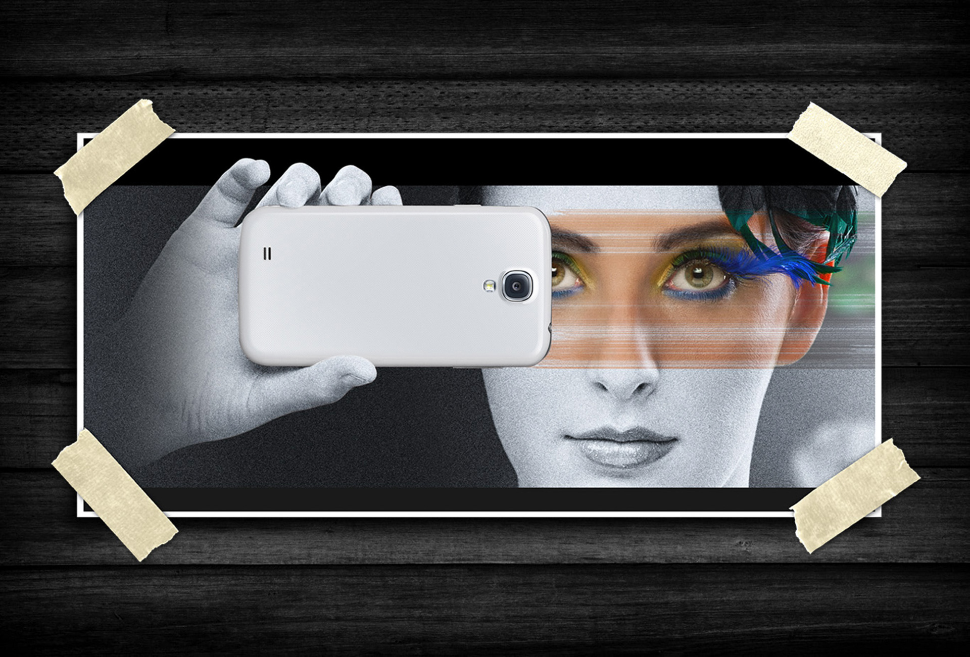 Creative portrait of a person holding a smartphone in front of their face, with selective color highlighting the eyes and vibrant feather-like makeup against a black-and-white background.