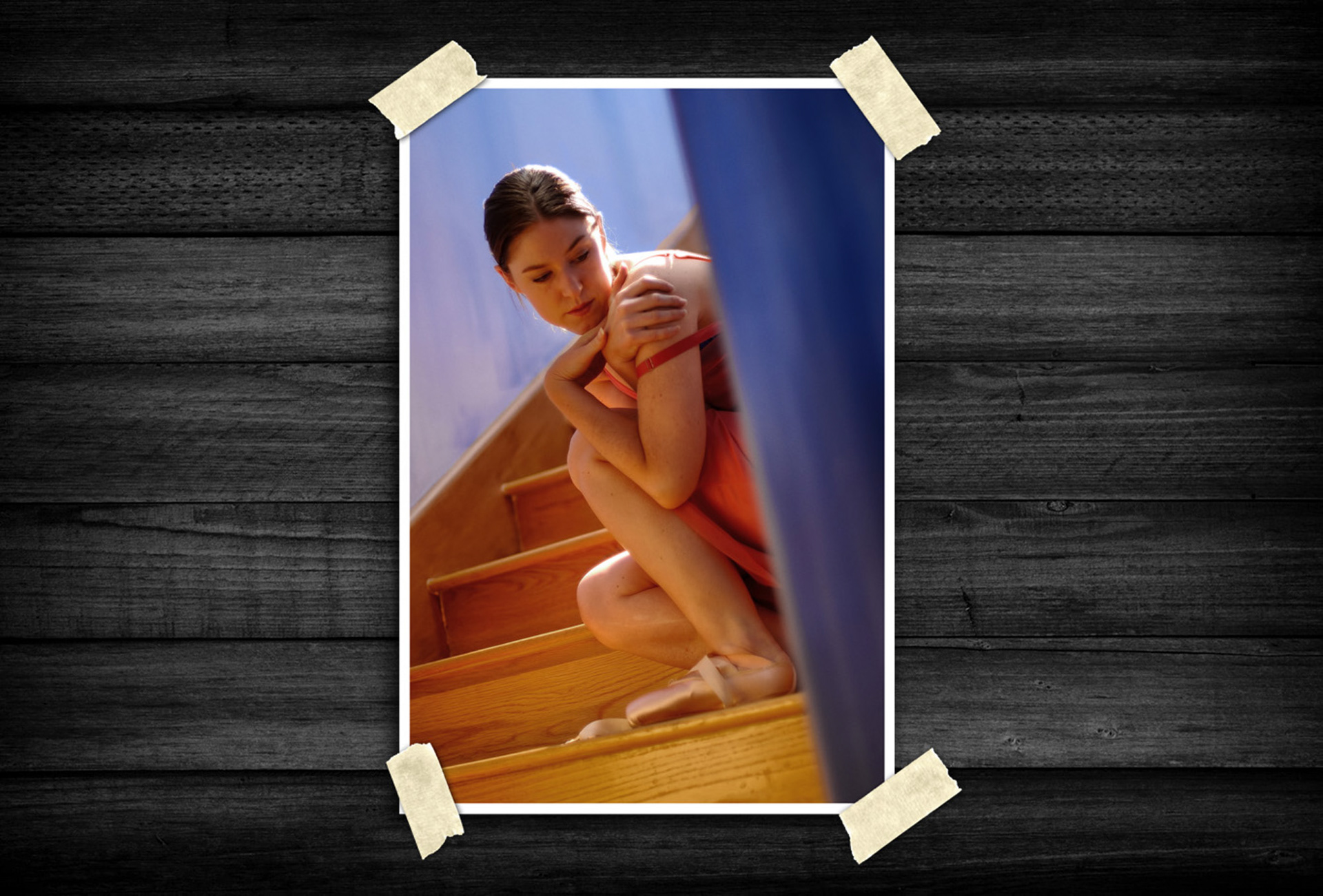 Portrait of a young ballet dancer sitting on a wooden staircase in pointe shoes, gently holding her shoulder in a quiet, contemplative pose.