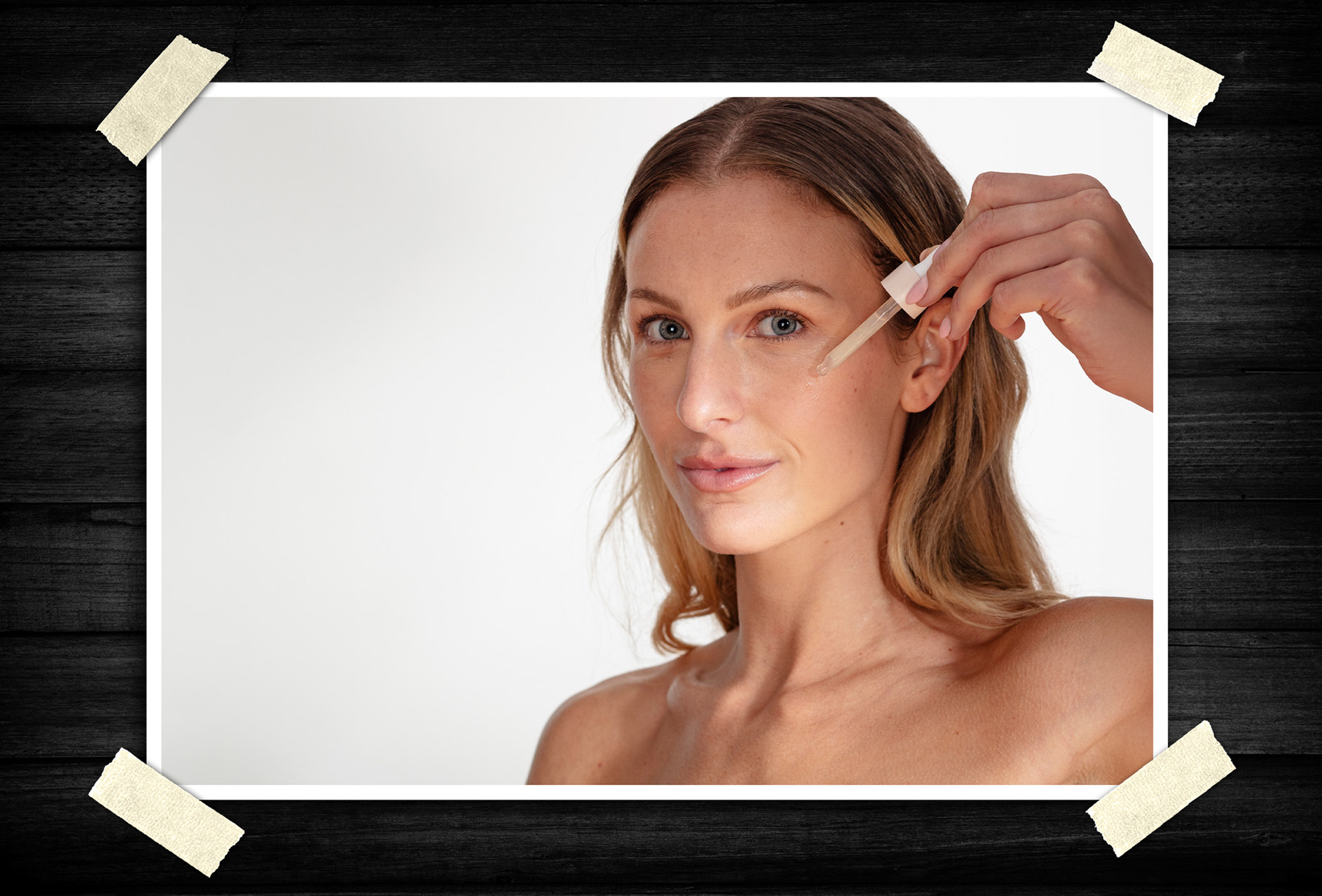 Clean beauty product portrait of a woman applying facial serum with a dropper while holding a skincare bottle against a light studio background.
