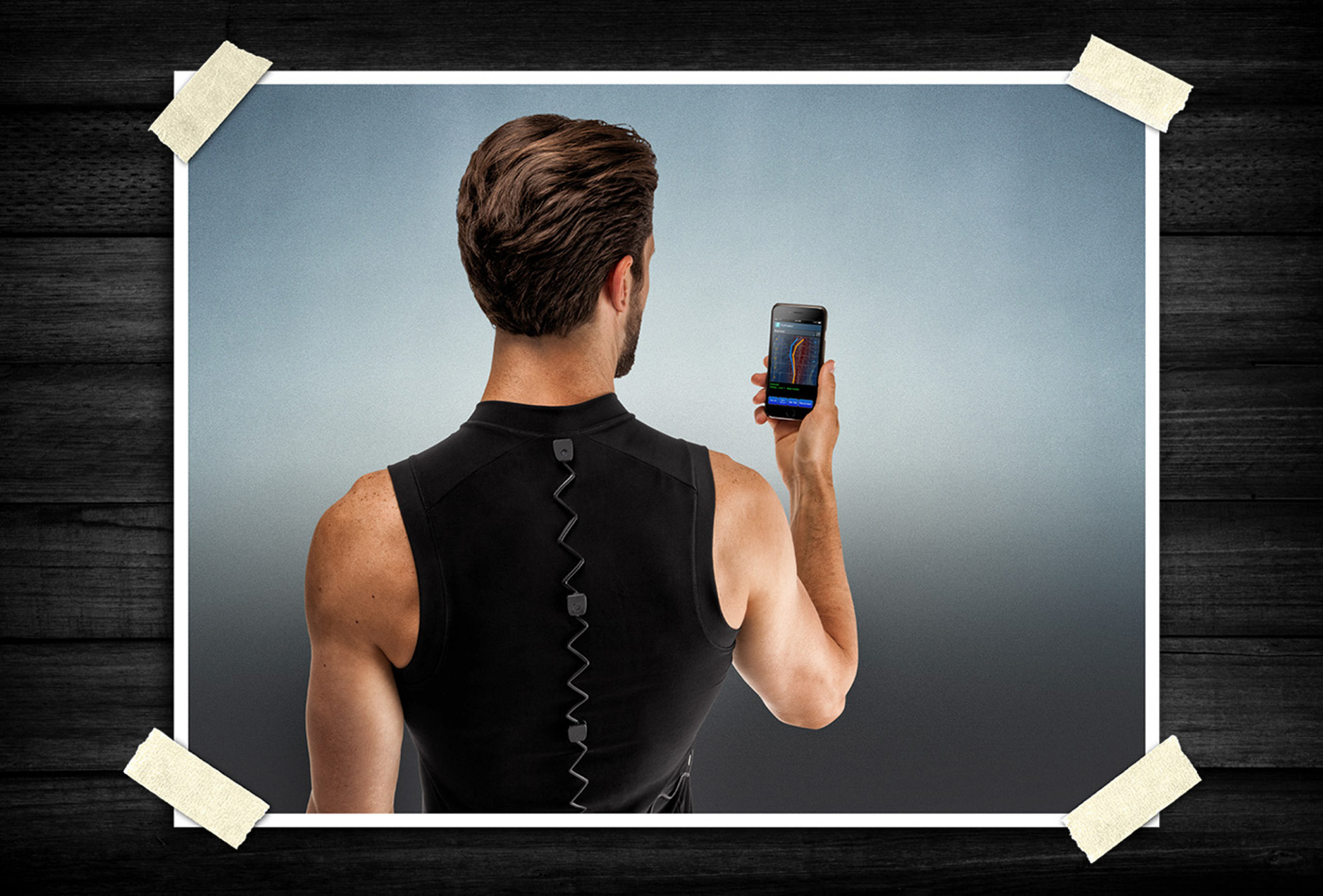 Rear view of a man wearing a smart fitness shirt with connected sensors while holding a smartphone displaying performance tracking data.
