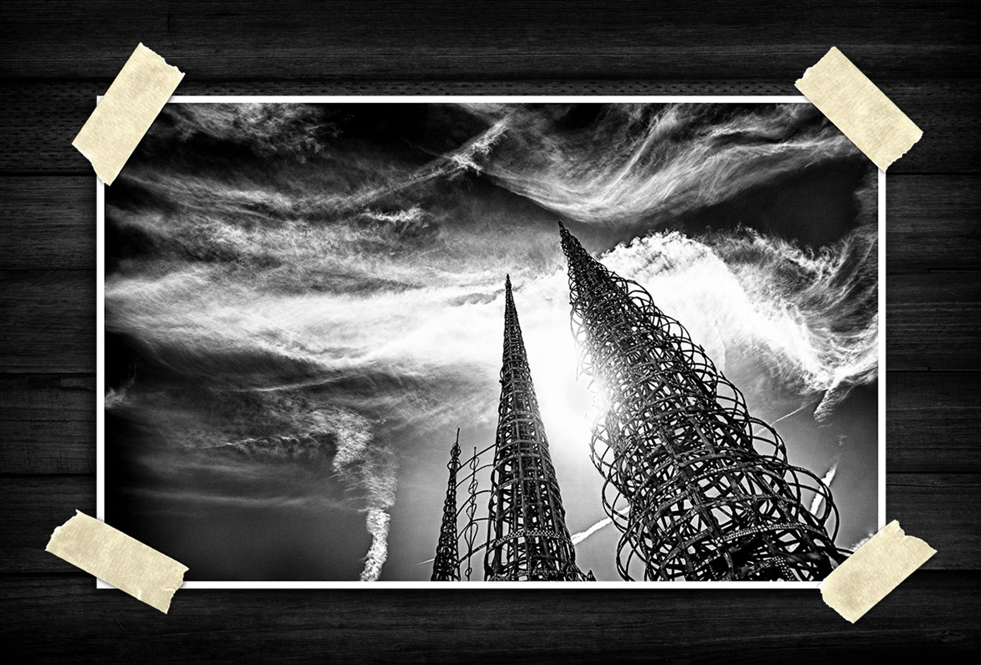 A dramatic black-and-white composition captures towering spiral metal structures rising sharply into the sky. Textured clouds sweep across the frame in layered patterns, while bright sunlight creates intense contrast and silhouettes, emphasizing height, scale, and architectural detail.