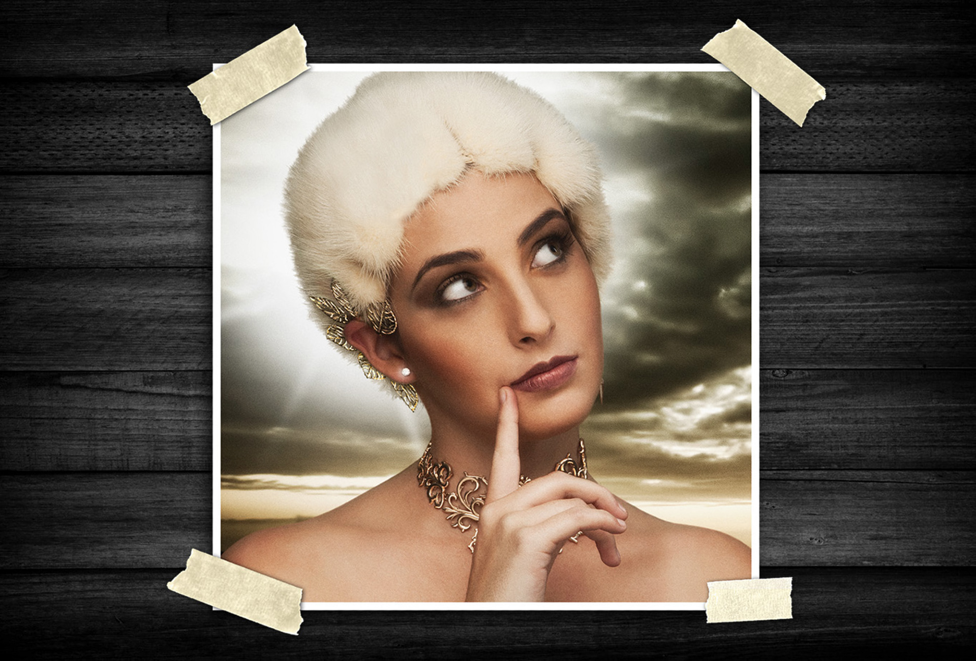 Close-up fashion portrait of a woman wearing a white fur hat with decorative gold jewelry, looking upward thoughtfully against a dramatic cloudy background.